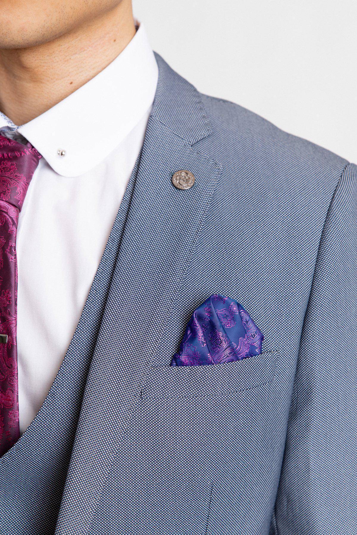 A close-up shows a person wearing the Marc Darcy DANNY - Blue Grey Tailored Blazer with a white shirt, purple paisley tie, and matching pocket square. Only the lower half of the face and upper torso are visible.