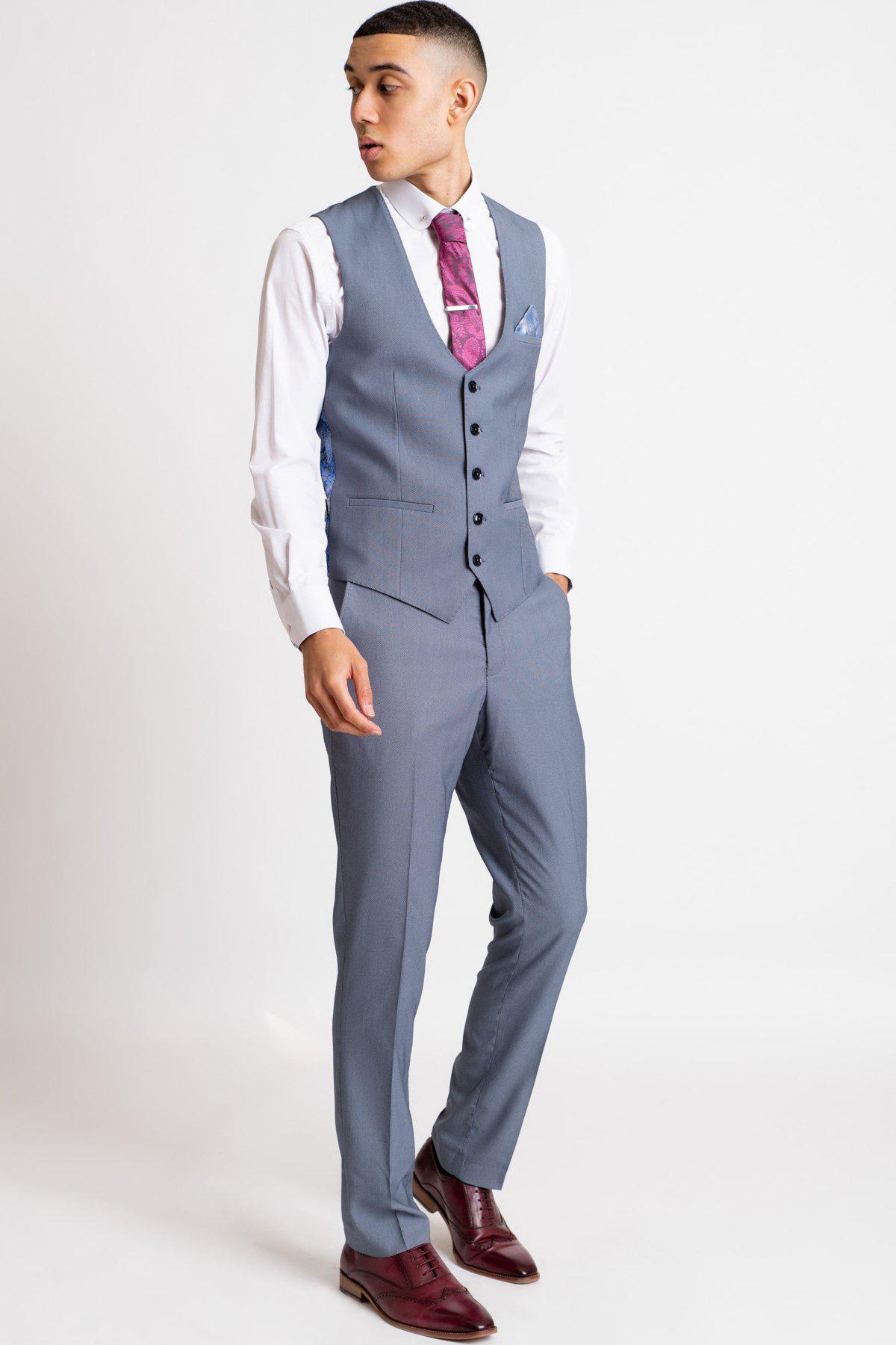 A man models Marc Darcy’s DANNY Blue Grey Tailored Trousers, styled with a white shirt, purple tie, and brown dress shoes, standing against a plain white background and looking left with one hand in his pocket.