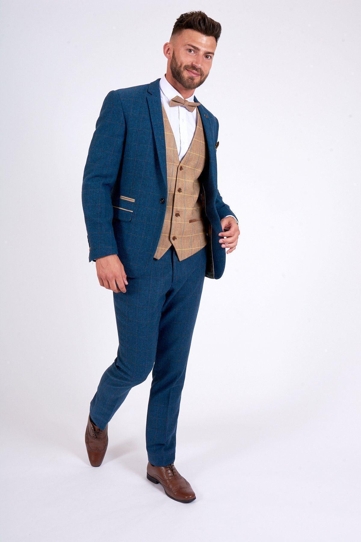 A man with short hair and a beard models the marcdarcy DION Blue Tweed Check Suit with DX7 Oak Waistcoat, white shirt, bow tie, and brown dress shoes. He stands mid-step, smiling against a plain white background.