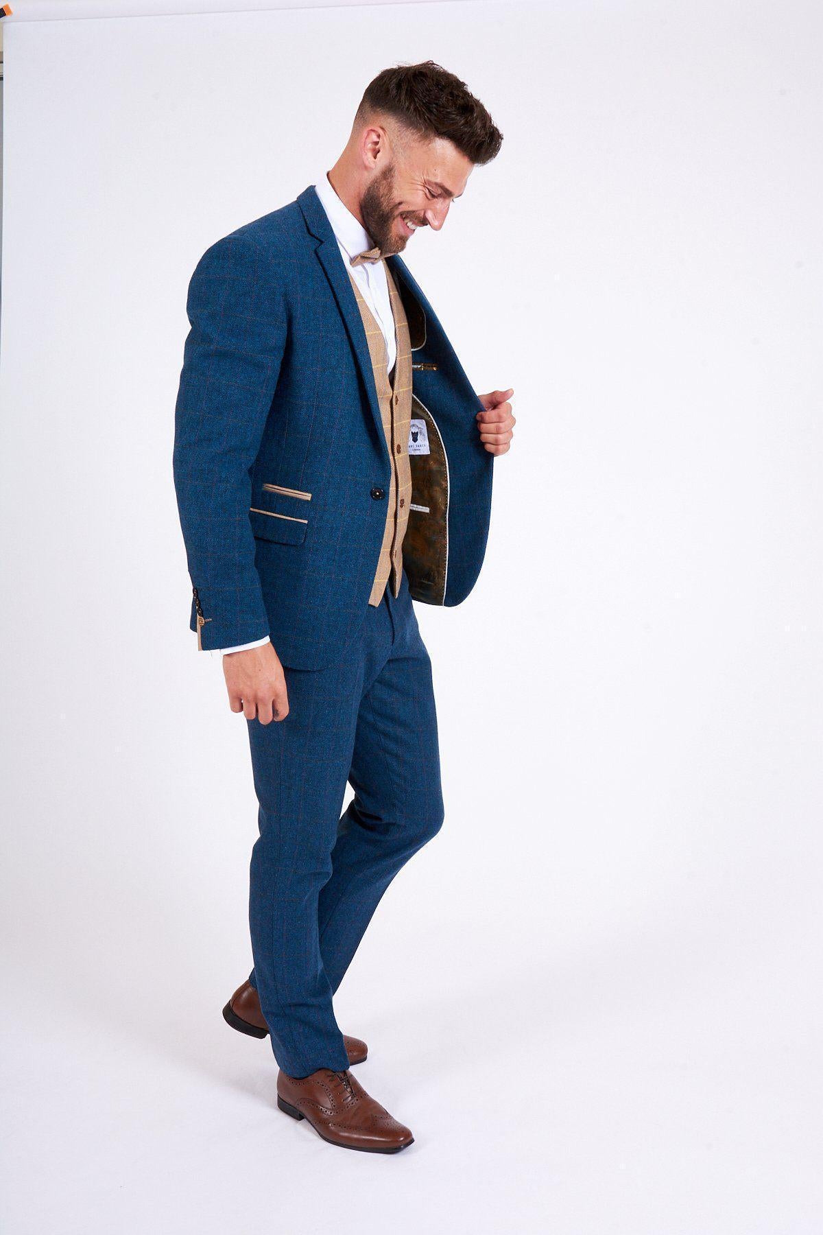 A man in the marcdarcy DION Blue Tweed Check Suit with DX7 Oak Waistcoat stands against a white background, smiling and looking down while holding his open jacket.