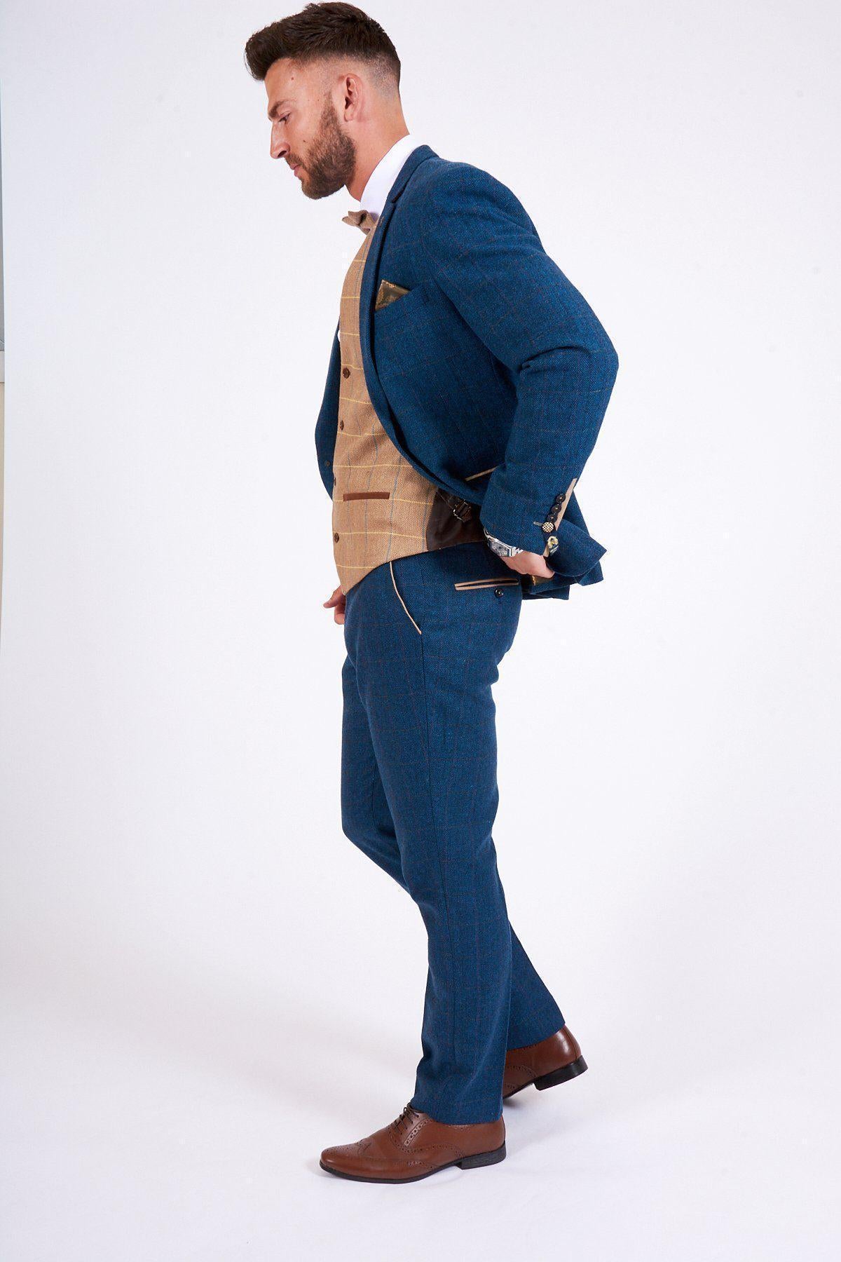 A man in the marcdarcy DION Blue Tweed Check Suit with DX7 Oak Waistcoat stands in profile against a white background, wearing a white shirt and brown shoes, holding a brown leather wallet behind his back.