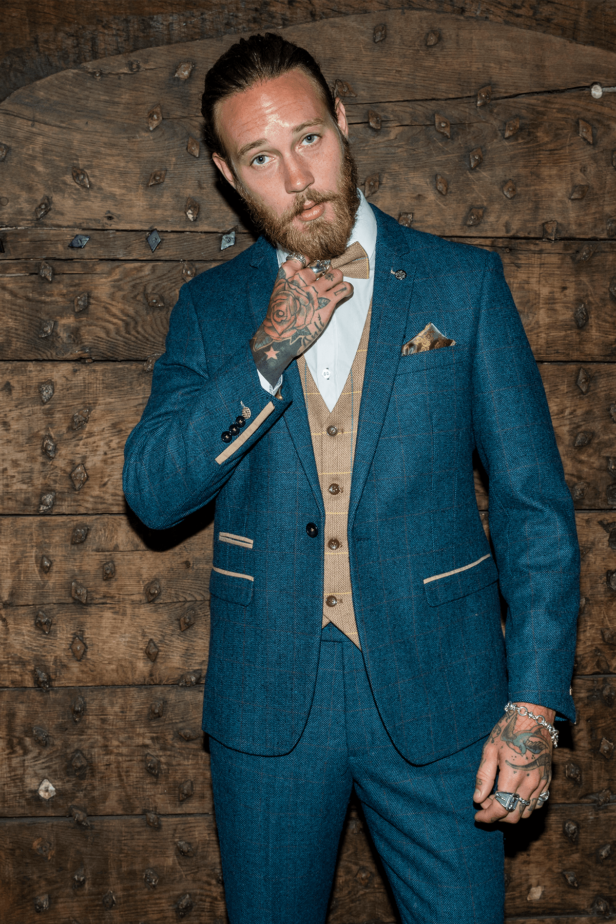 A tattooed, bearded man in the marcdarcy DION Blue Tweed Check Suit with DX7 Oak Waistcoat stands by a rustic wooden door, adjusting his tie and facing the camera.