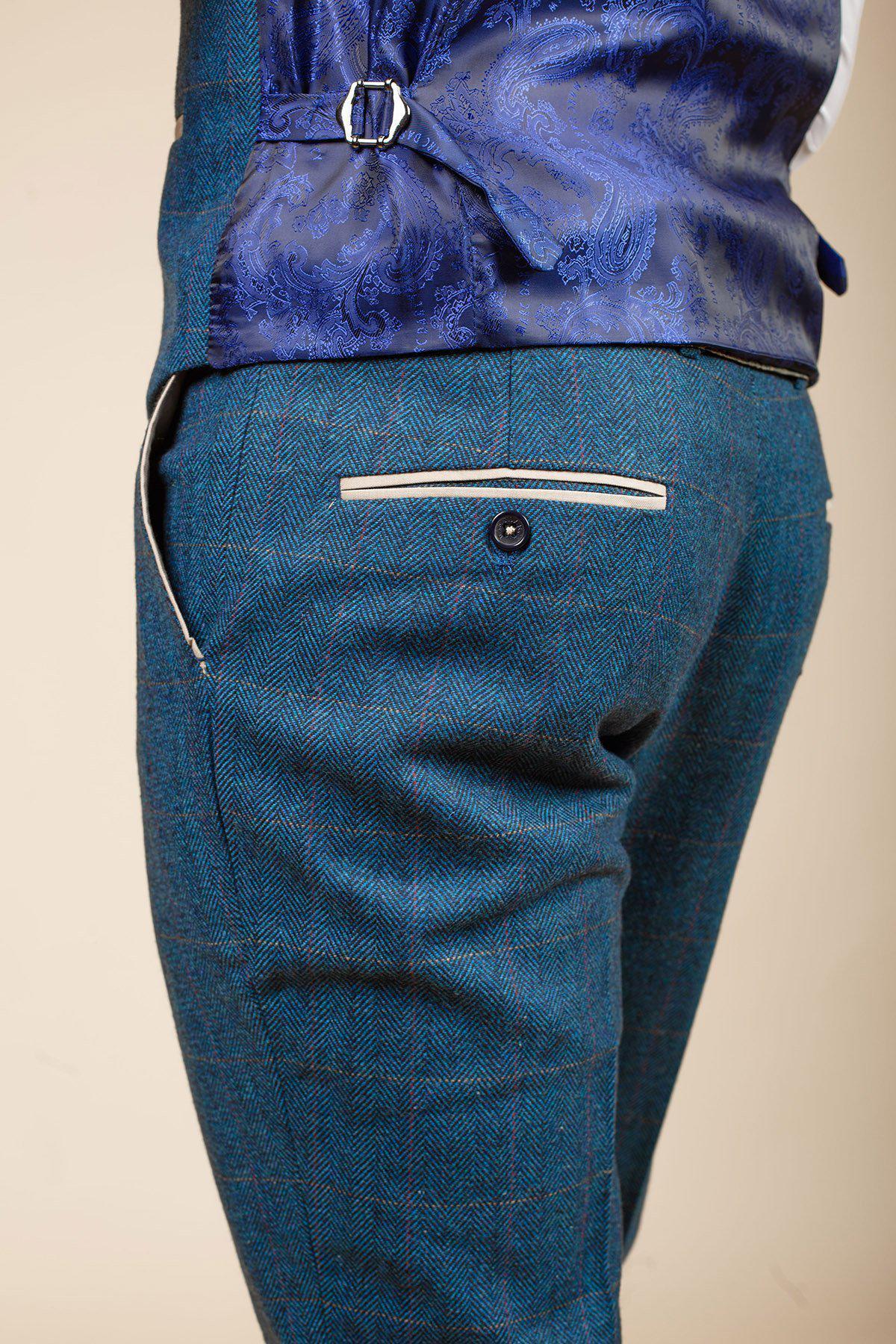 Close-up of a person wearing marcdarcys DION blue tweed check suit with Kelvin cream waistcoat, featuring teal herringbone trousers with a buttoned back pocket. Set against a plain beige background.
