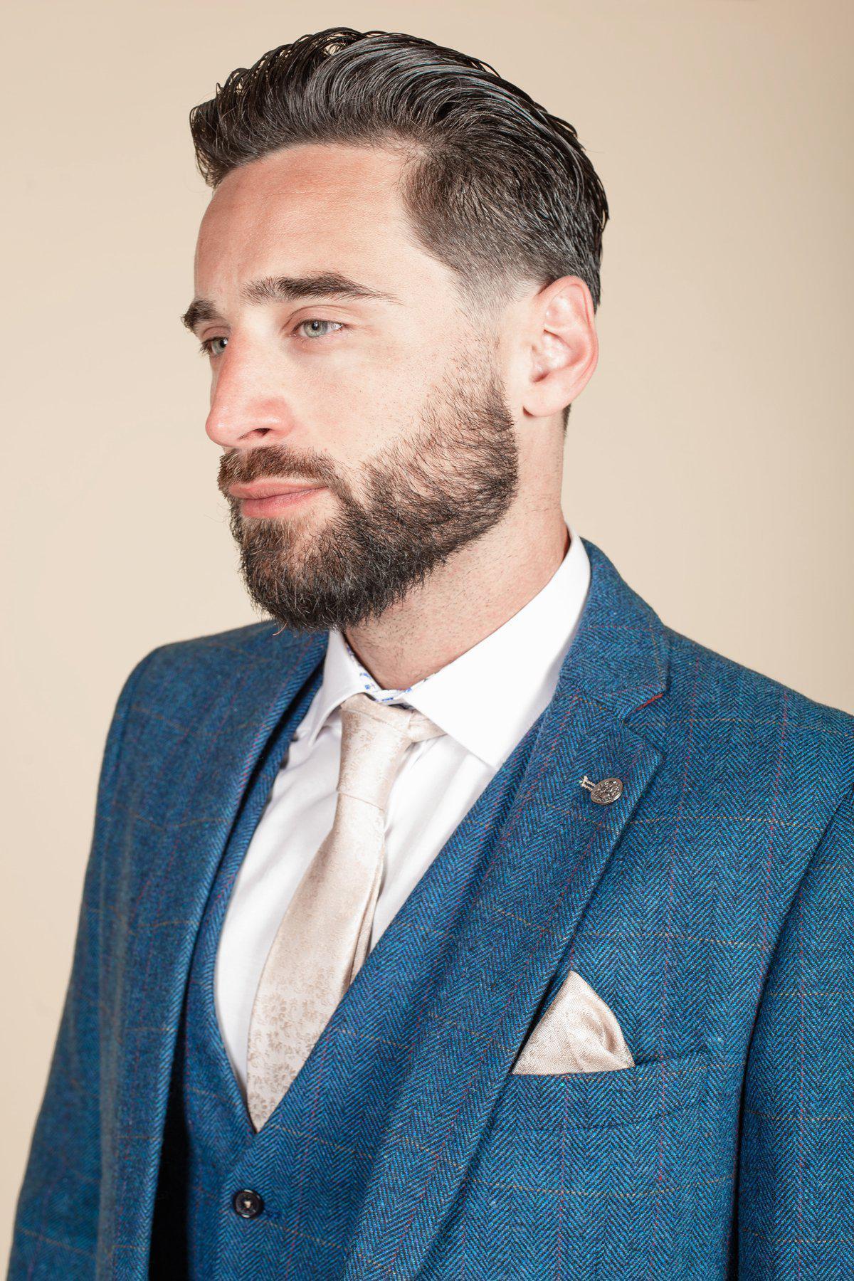 A man with slicked-back hair and a beard wears the marcdarcy DION Blue Tweed Check Suit with Kelvin Cream Waistcoat, styled with a white shirt and patterned tan tie—perfect as a groom suit or stylish wedding suit.