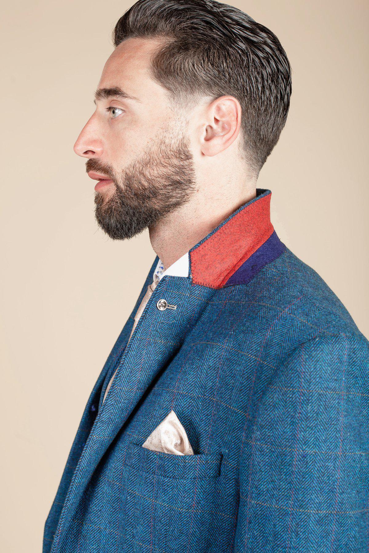 A bearded man with slicked-back hair wears the marcdarcy DION blue tweed check suit with a Kelvin cream waistcoat, white shirt, and cream pocket square, shown in profile against a beige background.