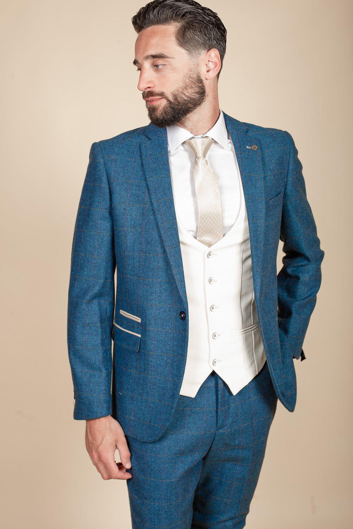 A man with dark hair and a beard wears the marcdarcy DION blue tweed check suit with Kelvin cream waistcoat, paired with a white shirt and cream tie. He looks left, one hand in his pocket, against a plain beige background.