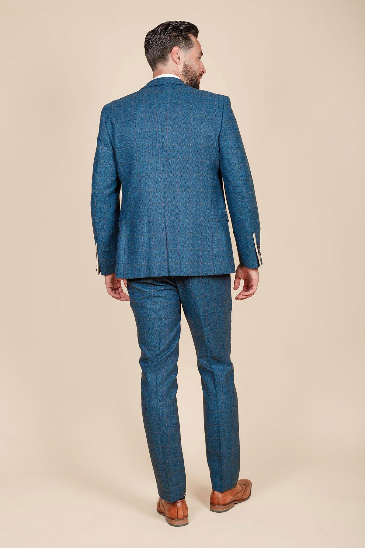 A man faces away, wearing the marcdarcy DION blue tweed check suit with a Kelvin cream waistcoat and brown dress shoes, set against a plain beige background.