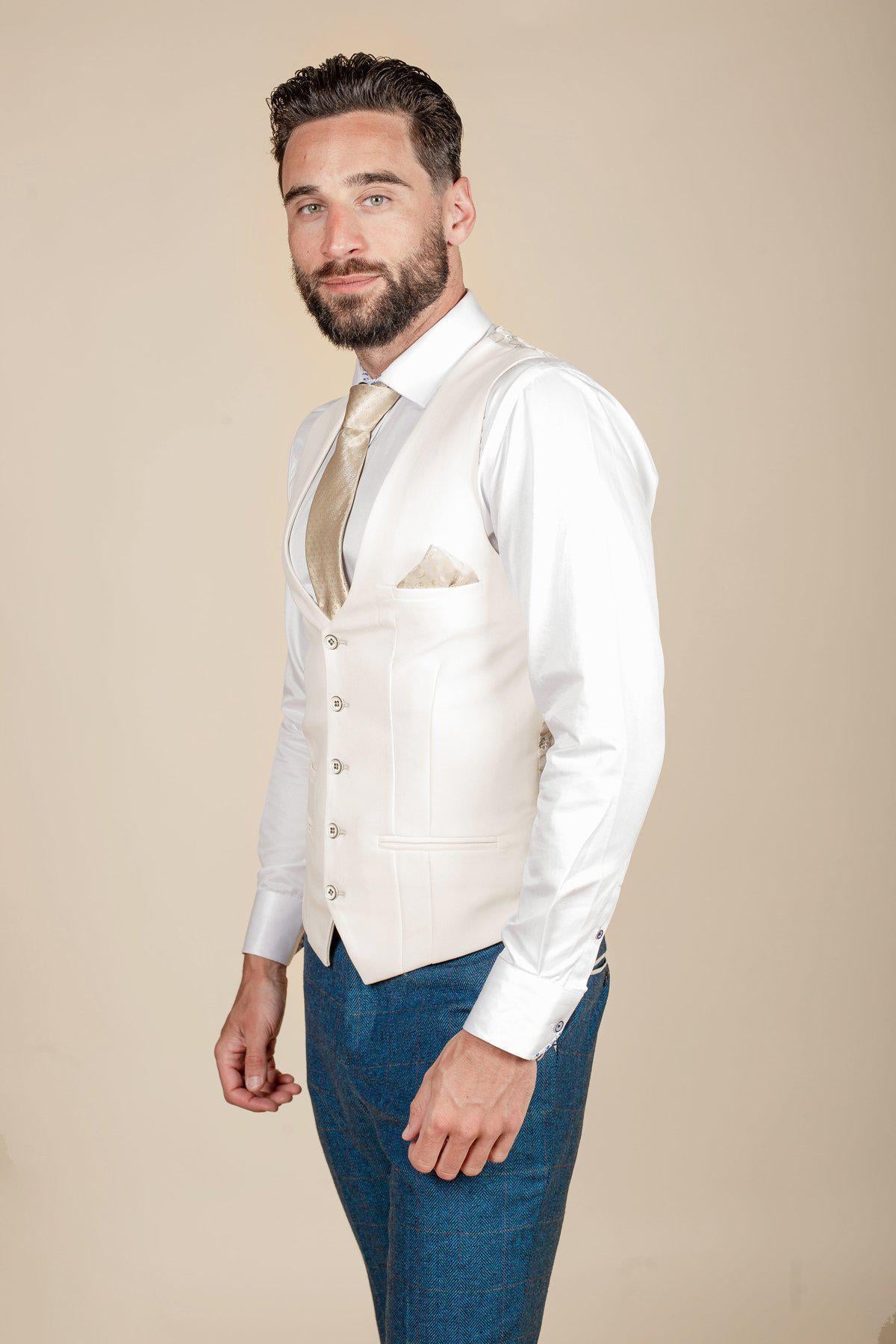 A man with dark hair and a beard wears the marcdarcy DION Blue Tweed Check Suit with the Kelvin Cream Waistcoat over a white shirt, gold tie, and pocket square, paired with blue pants, against a beige background, smiling slightly.