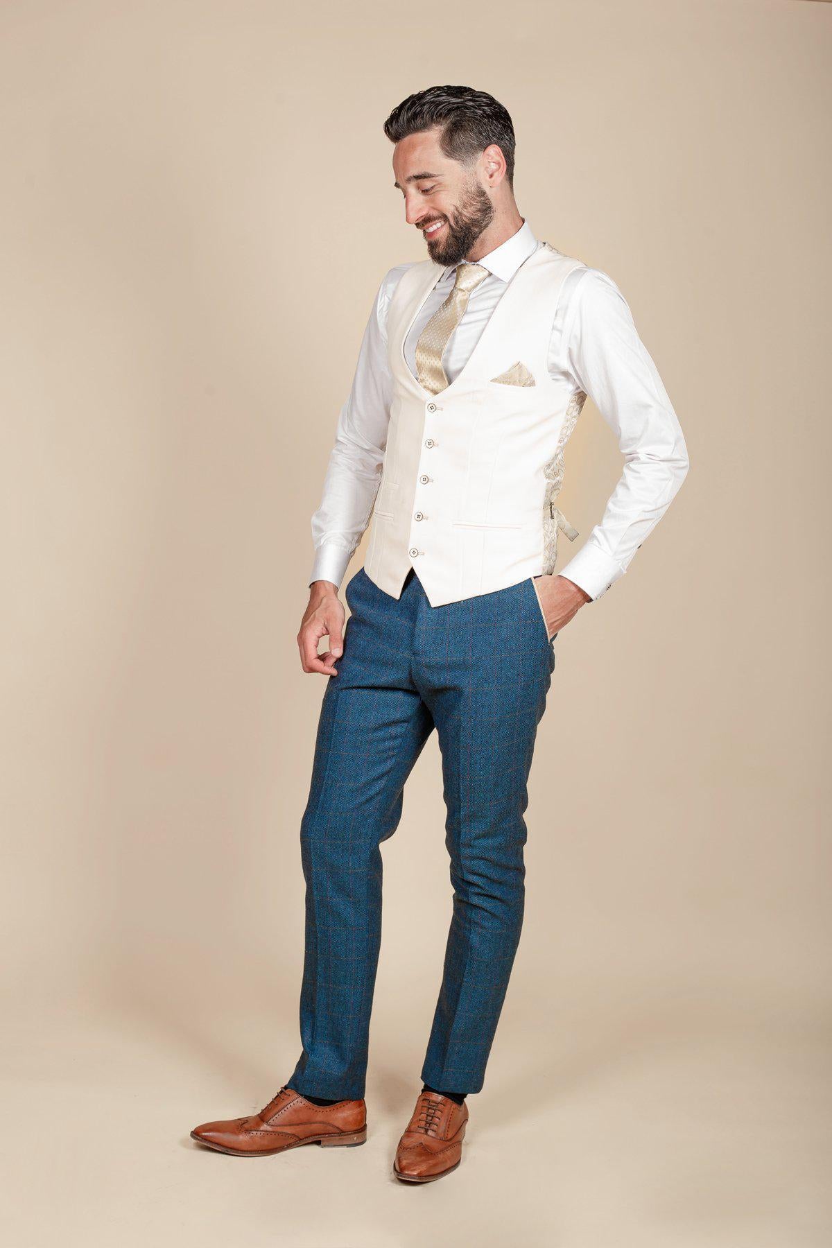 A bearded man smiles against a beige background, wearing the marcdarcy DION Blue Tweed Check Suit with a Kelvin cream waistcoat, white shirt, gold tie, blue trousers, and brown dress shoes—perfectly styled for a wedding.
