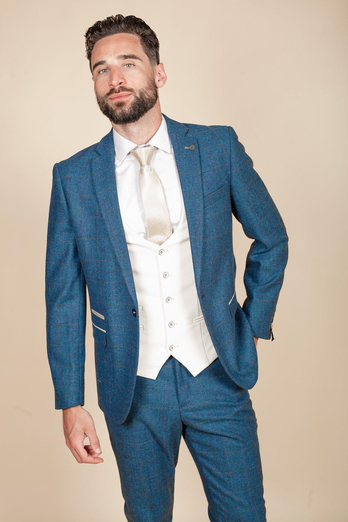A man with dark hair and a beard models the marcdarcy DION blue tweed check suit with a Kelvin cream waistcoat, white dress shirt, and cream tie, standing against a beige background with one hand in his pocket.
