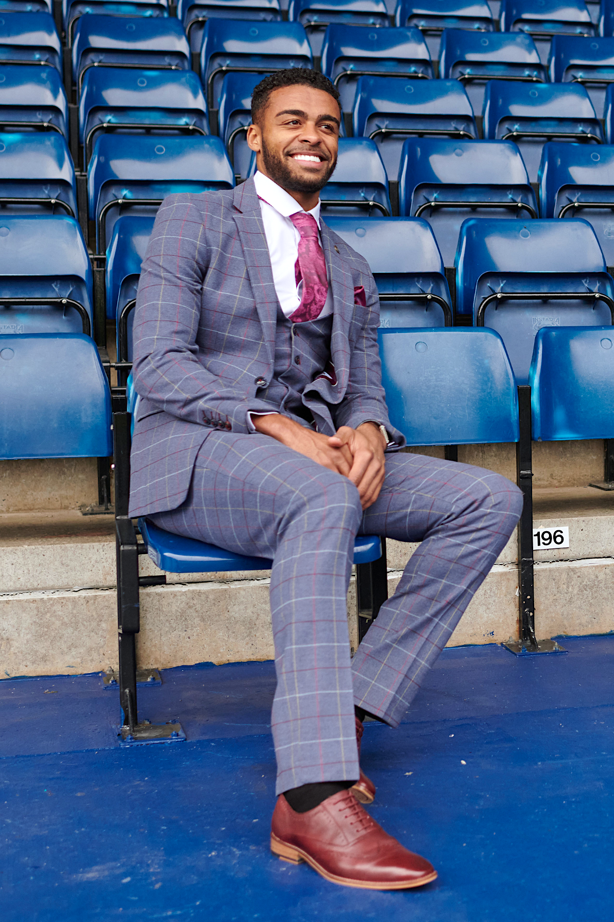 A man in the marcdarcy WBA Collection - Drake Sky Suit as worn by Darnell Furlong, paired with a red tie and brown dress shoes, sits smiling on blue stadium seats with legs crossed and hands on his knee.
