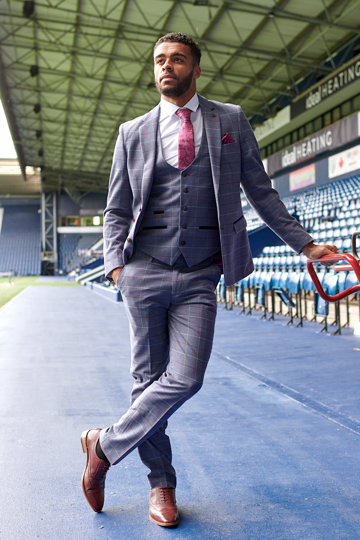 Wearing marcdarcy’s The WBA Collection - Drake Sky Suit as worn by Darnell Furlong, a man in a slim-fit check suit and pink tie stands confidently by the red rail, stadium blue seats and metal roof behind him—a real marcdarcy style moment.