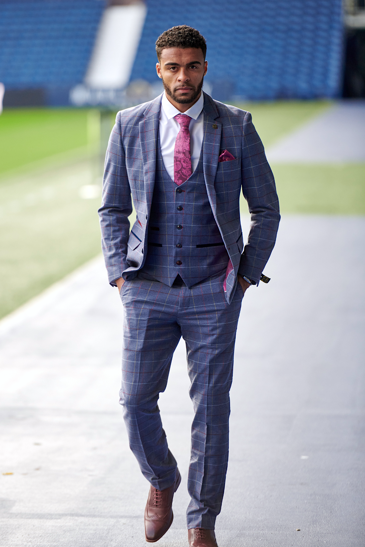 A man in the marcdarcy WBA Collection Drake Sky Suit, as worn by Darnell Furlong, paired with a pink tie, walks confidently beside a sports field with empty stadium seats in the background, hands in pockets.