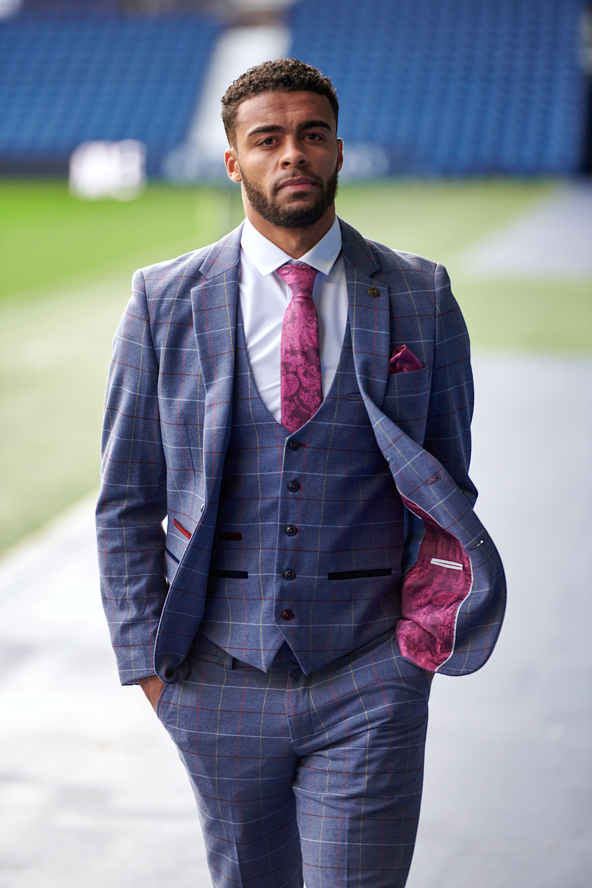 A man walks confidently near a sports stadium in the marcdarcy WBA Collection - Drake Sky Suit as worn by Darnell Furlong, featuring a slim fit blue check and pink tie, with blurred blue seats in the background.
