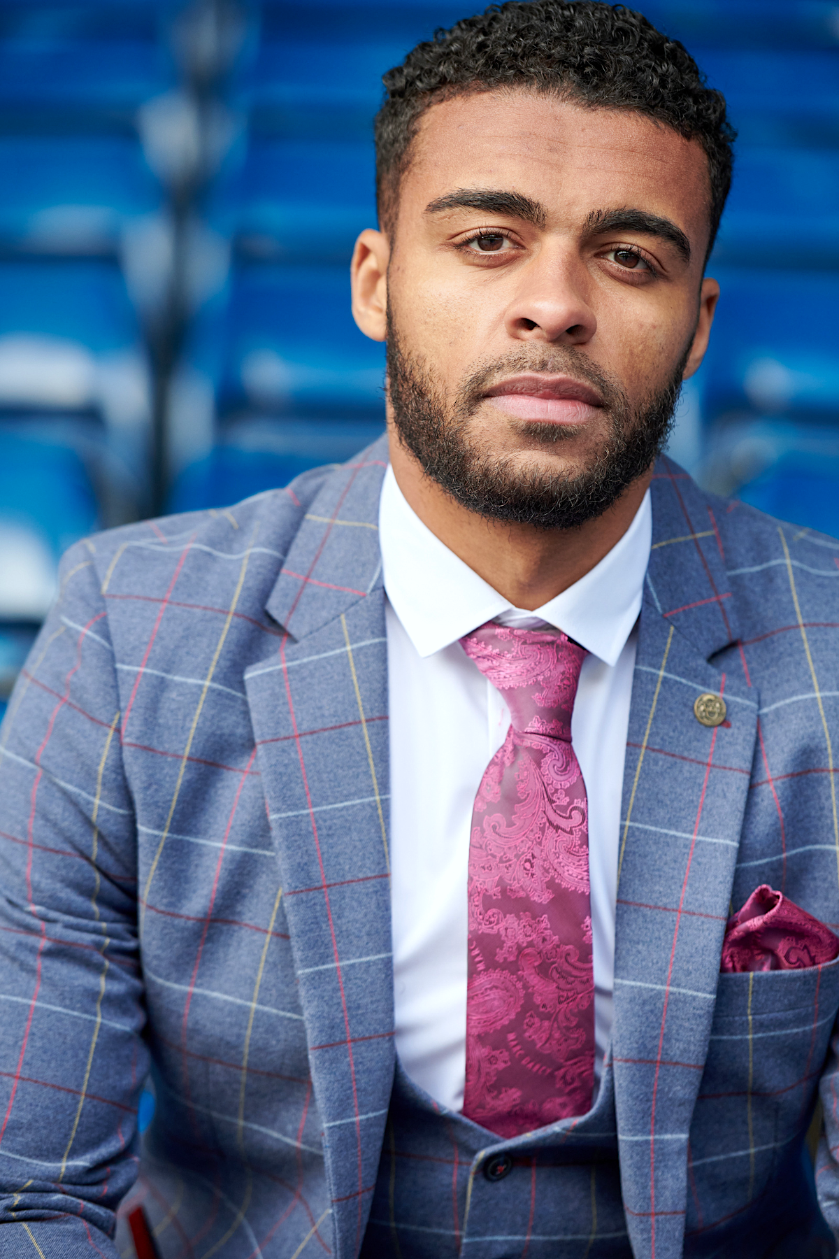 Close-up of a man with dark, curly hair and a beard wearing the marcdarcy WBA Collection - Drake Sky Suit as worn by Darnell Furlong: a grey checked slim fit suit paired with a pink patterned tie and pocket square.
