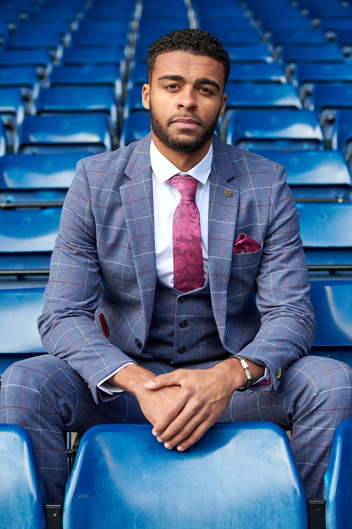 A man in the marcdarcy WBA Collection - Drake Sky Suit, as worn by Darnell Furlong, with a pink tie and pocket square sits on blue stadium seats, gazing seriously at the camera.