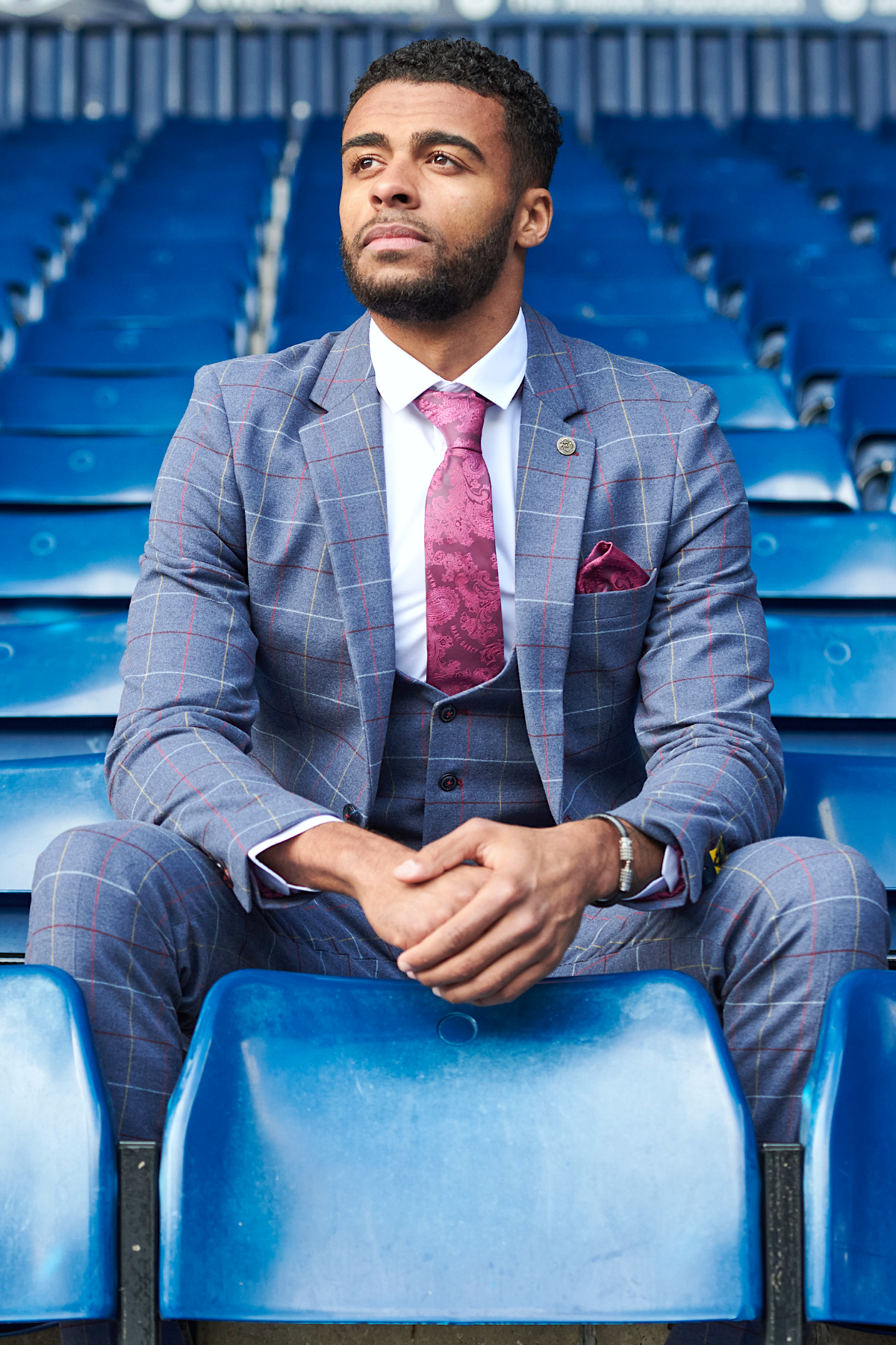 A man wearing the marcdarcy WBA Collection - Drake Sky Suit as worn by Darnell Furlong, paired with a white shirt and pink patterned tie, sits alone in blue stadium seats, gazing thoughtfully into the distance.