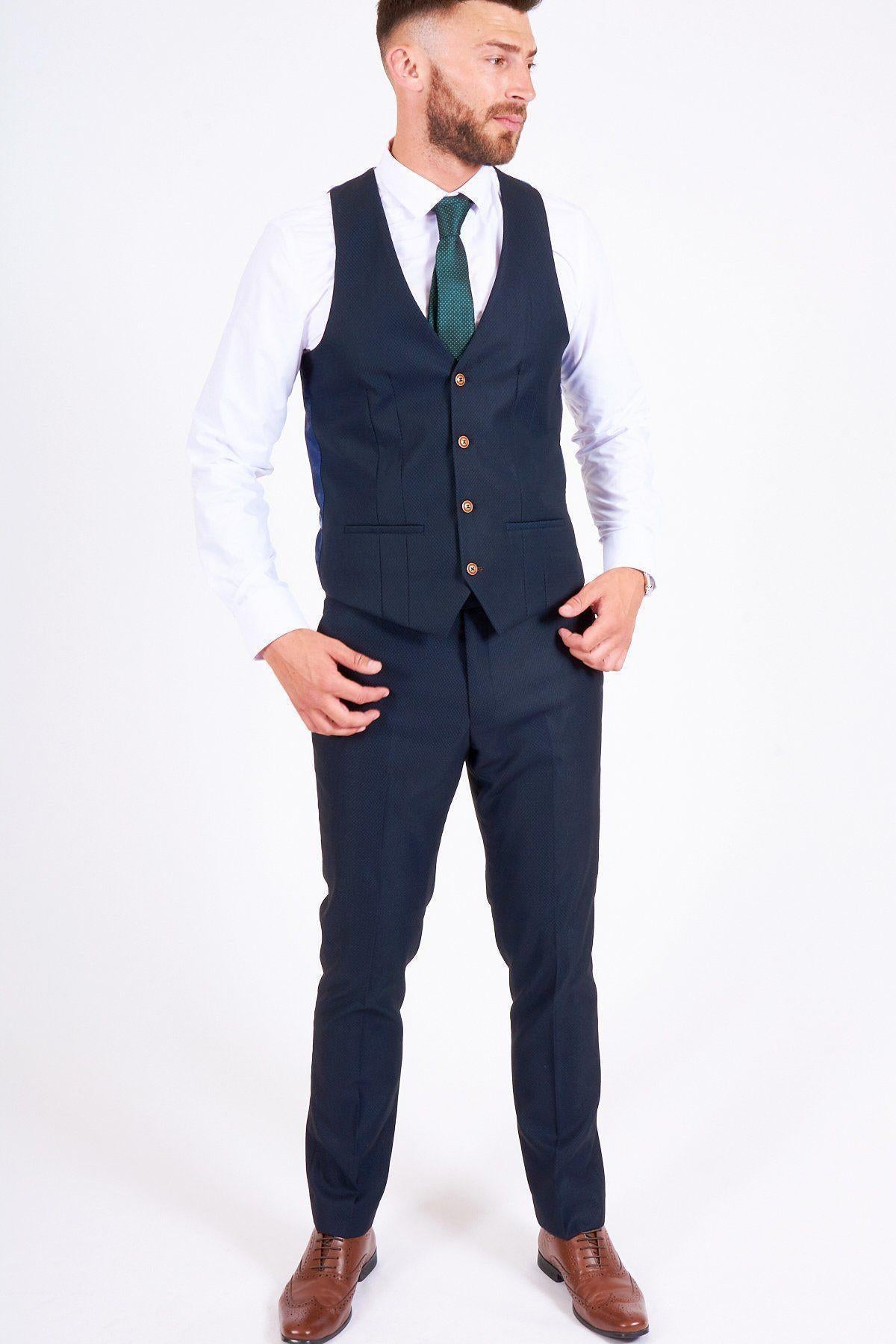 James Bye, as Martin Fowler, wears the marcdarcy Max Navy Suit: a slim fit navy blazer and trousers with a white shirt, green tie, and brown shoes. He stands against a white background with short hair and a trimmed beard, hands by his waist.