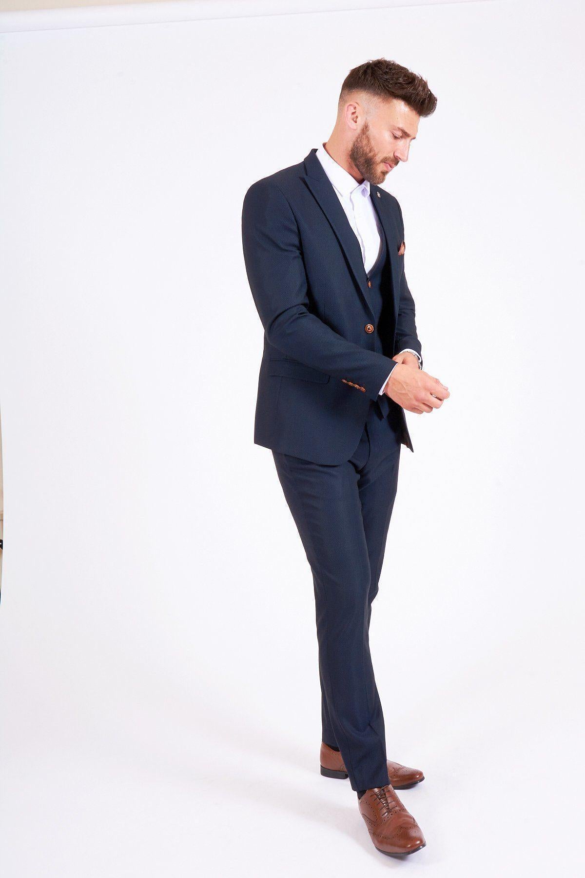 James Bye as Eastenders Martin Fowler wears the marcdarcy Max Navy slim fit suit with a white shirt and brown shoes, standing against a white background, adjusting his cufflink while looking down and slightly smiling.