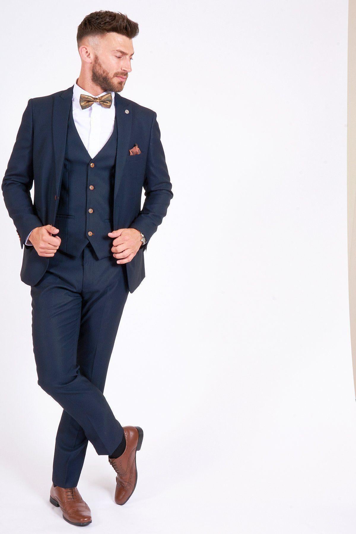 James Bye, as Eastenders' Martin Fowler, wears the marcdarcy Max Navy Suit—a dark navy slim fit single-breasted blazer with a white shirt and brown bow tie—posing with crossed legs against a white background.