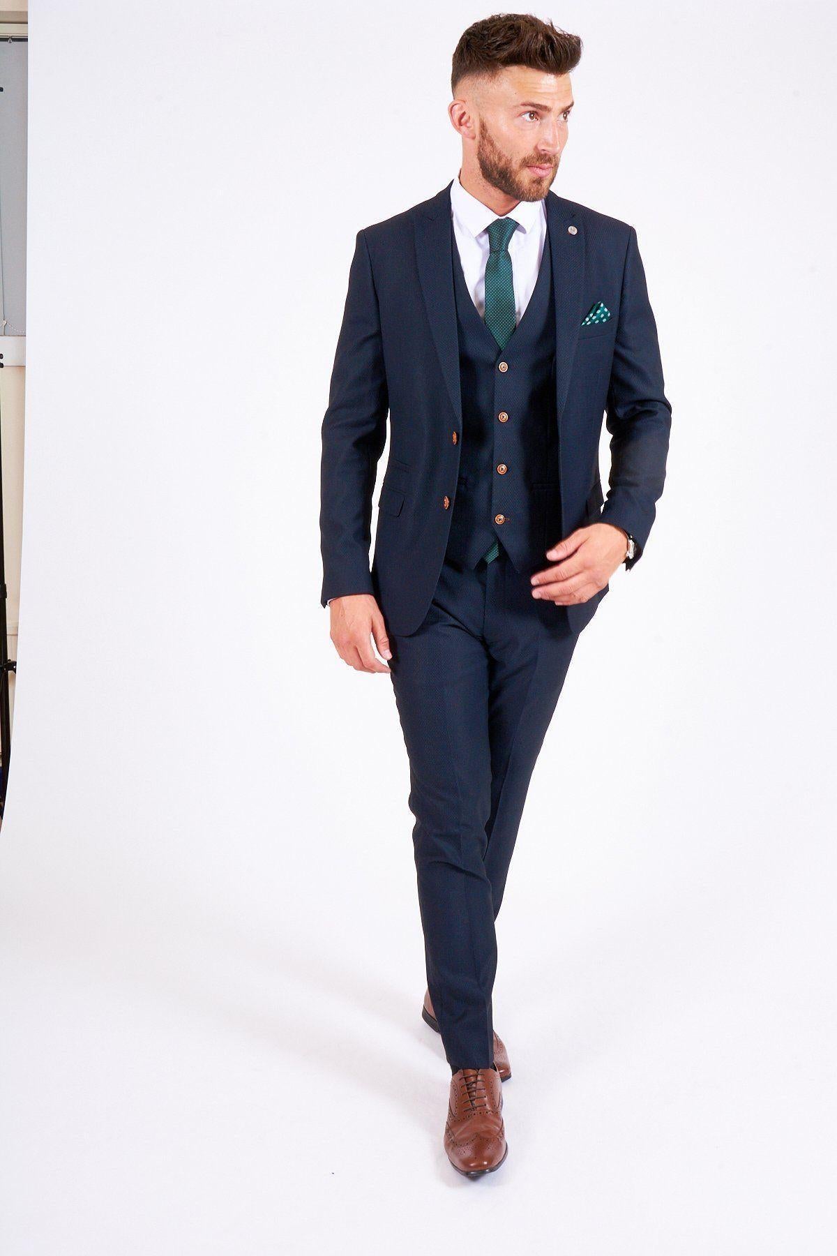 James Bye as Eastenders’ Martin Fowler wears the marcdarcy Max Navy Slim Fit Three-Piece Suit with a white shirt, green tie, and brown shoes, standing against a plain white background with one hand in his pocket and looking to the side.