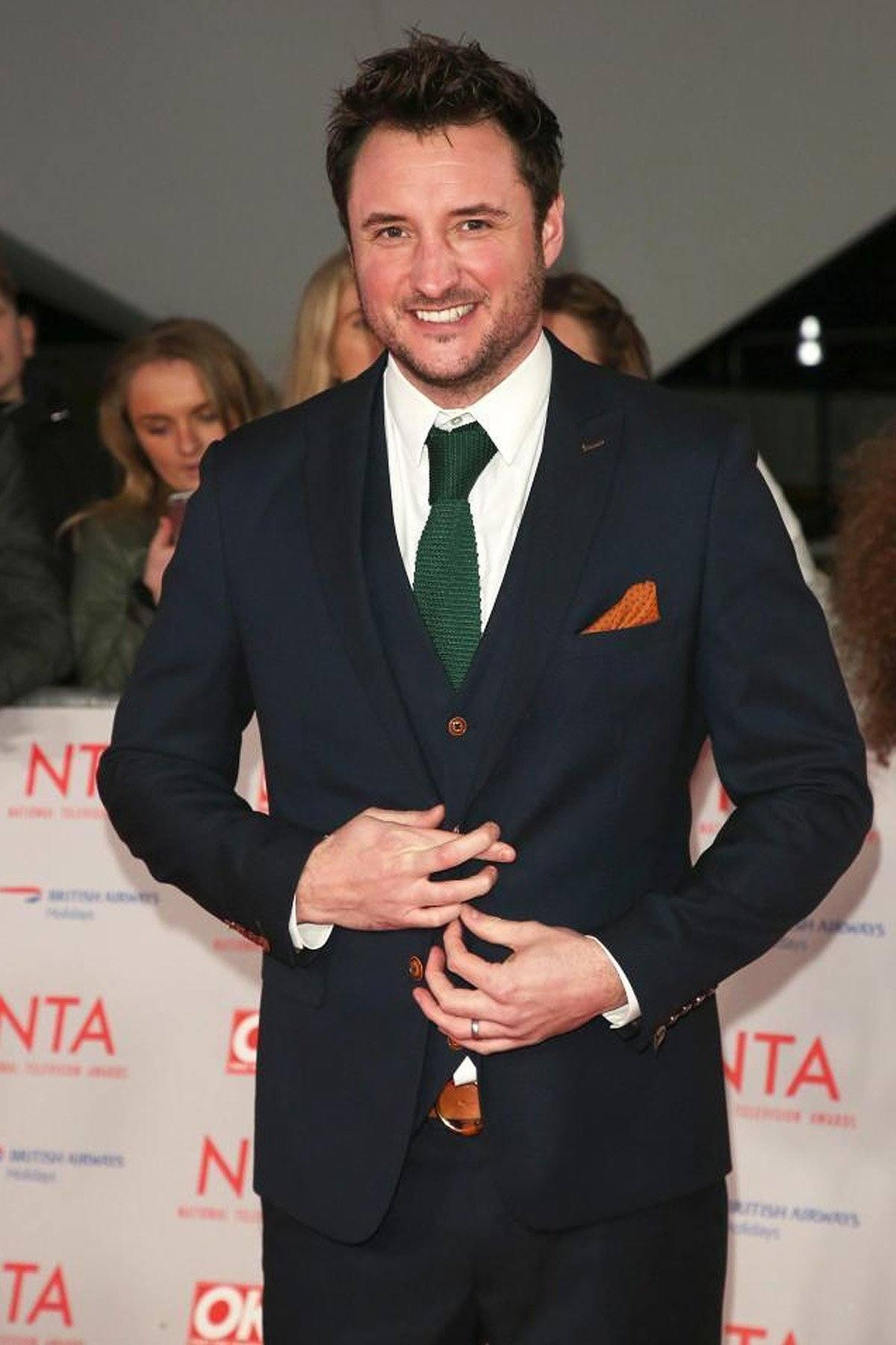 Eastenders Martin Fowler (James Bye) wears a marcdarcy Max navy suit, styled with a green tie and orange pocket square, as he smiles and poses on the red carpet at an event.
