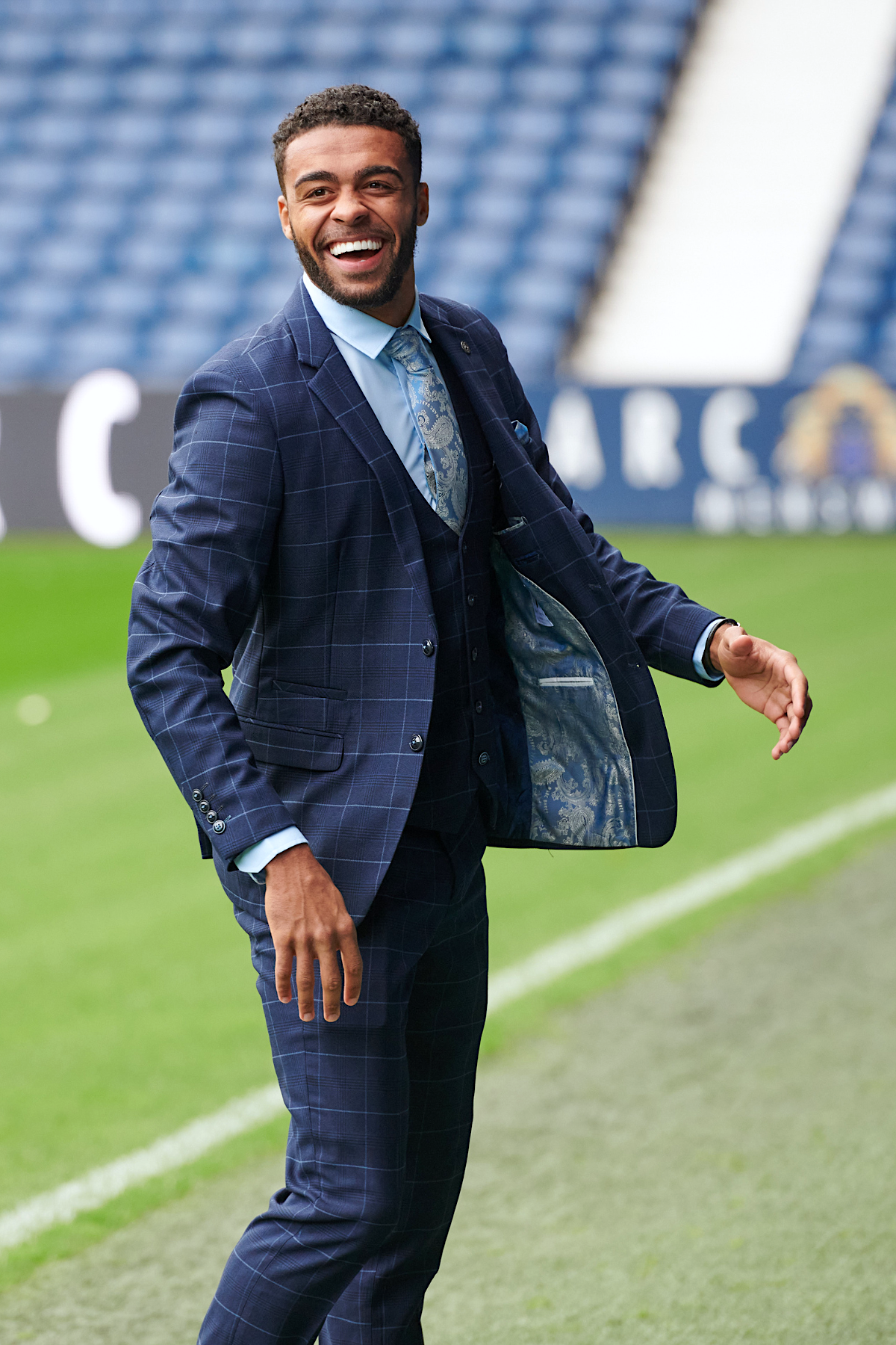 A Black man in the marcdarcy WBA Collection Edinson Navy & Sky Check Suit, paired with a light blue shirt and patterned tie, smiles widely at a sporting event.