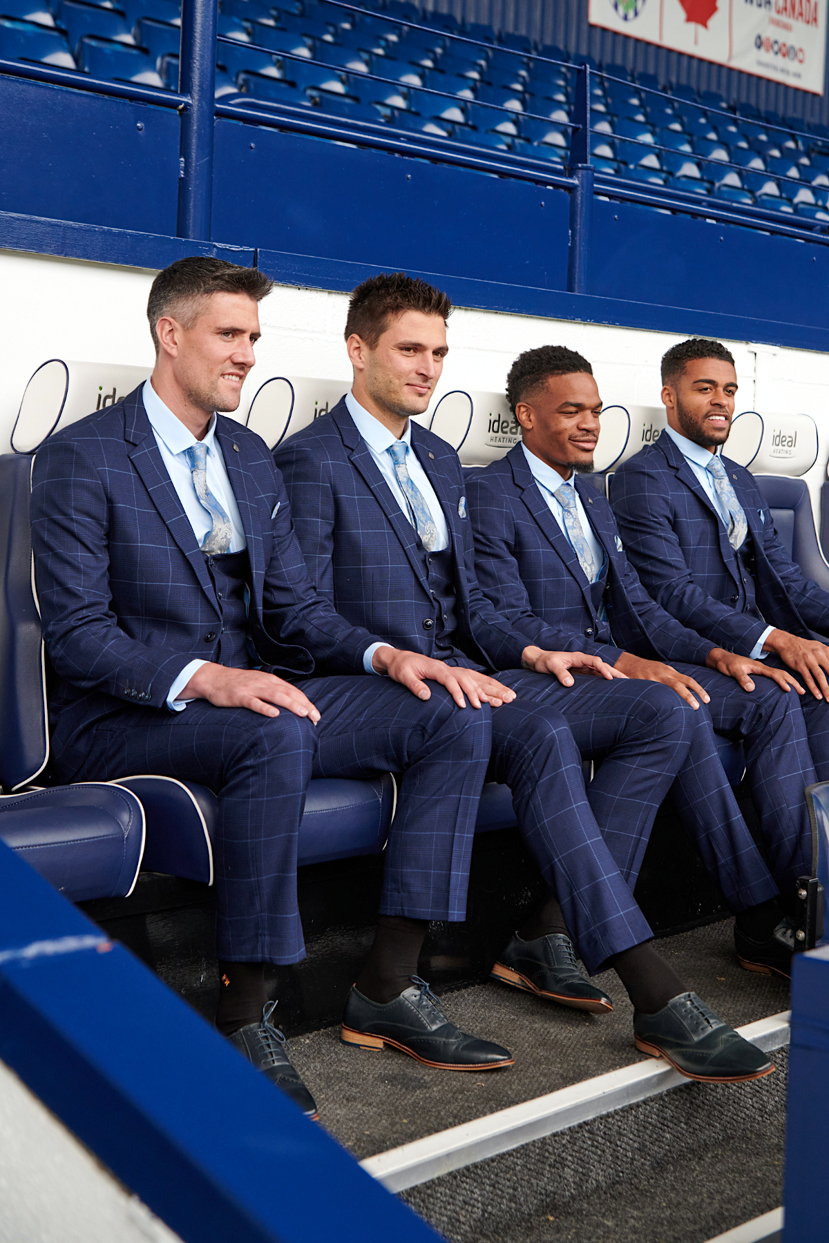Four men wear marcdarcy’s WBA Collection Edinson Navy & Sky Check three-piece suits with light blue shirts and ties, smiling as they sit on a stadium bench in an empty sports venue.