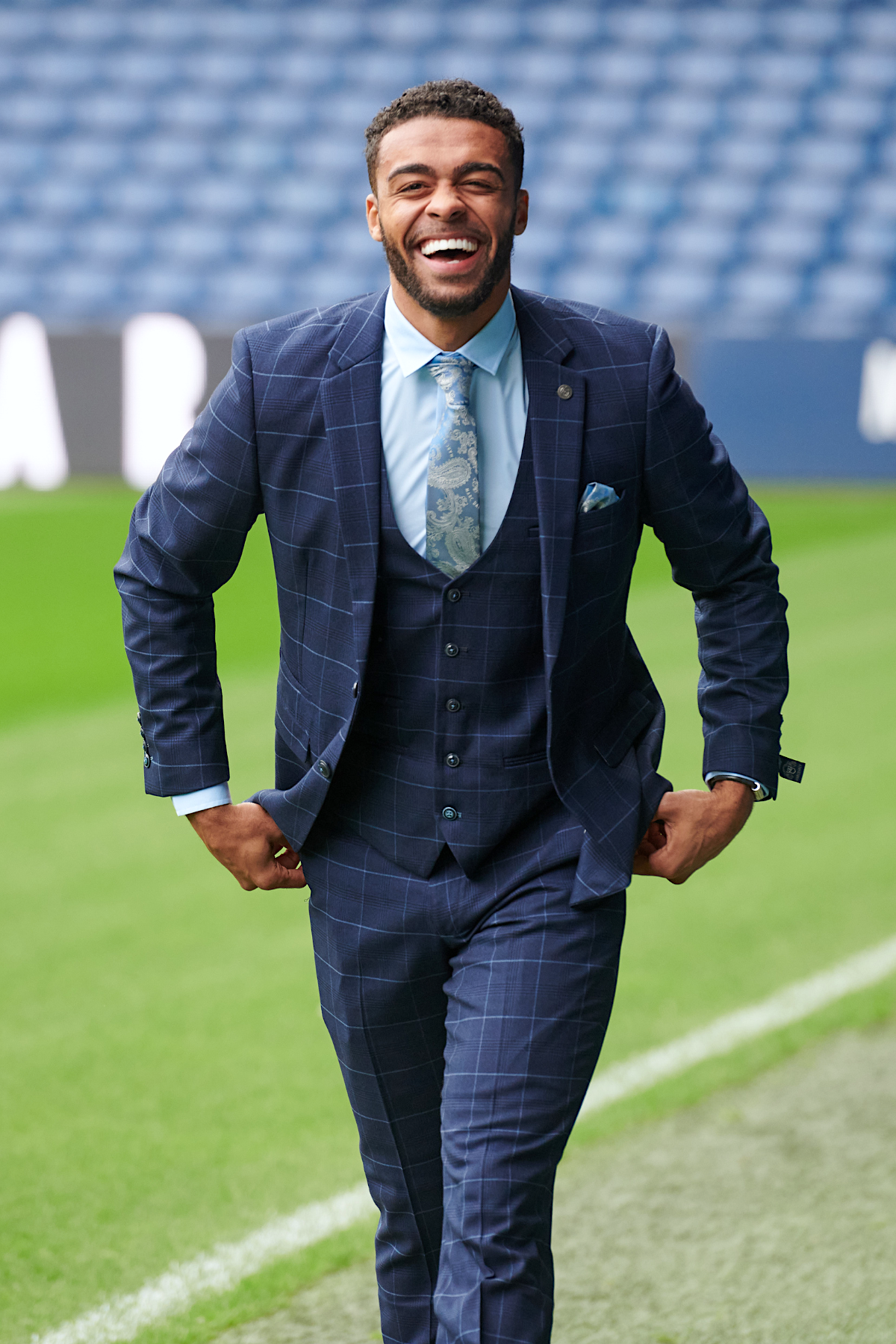 A man in the marcdarcy Edinson Navy & Sky Check Suit from The WBA Collection smiles as he walks on a sports field with West Bromwich Albion F.C. stadium seating in the background.