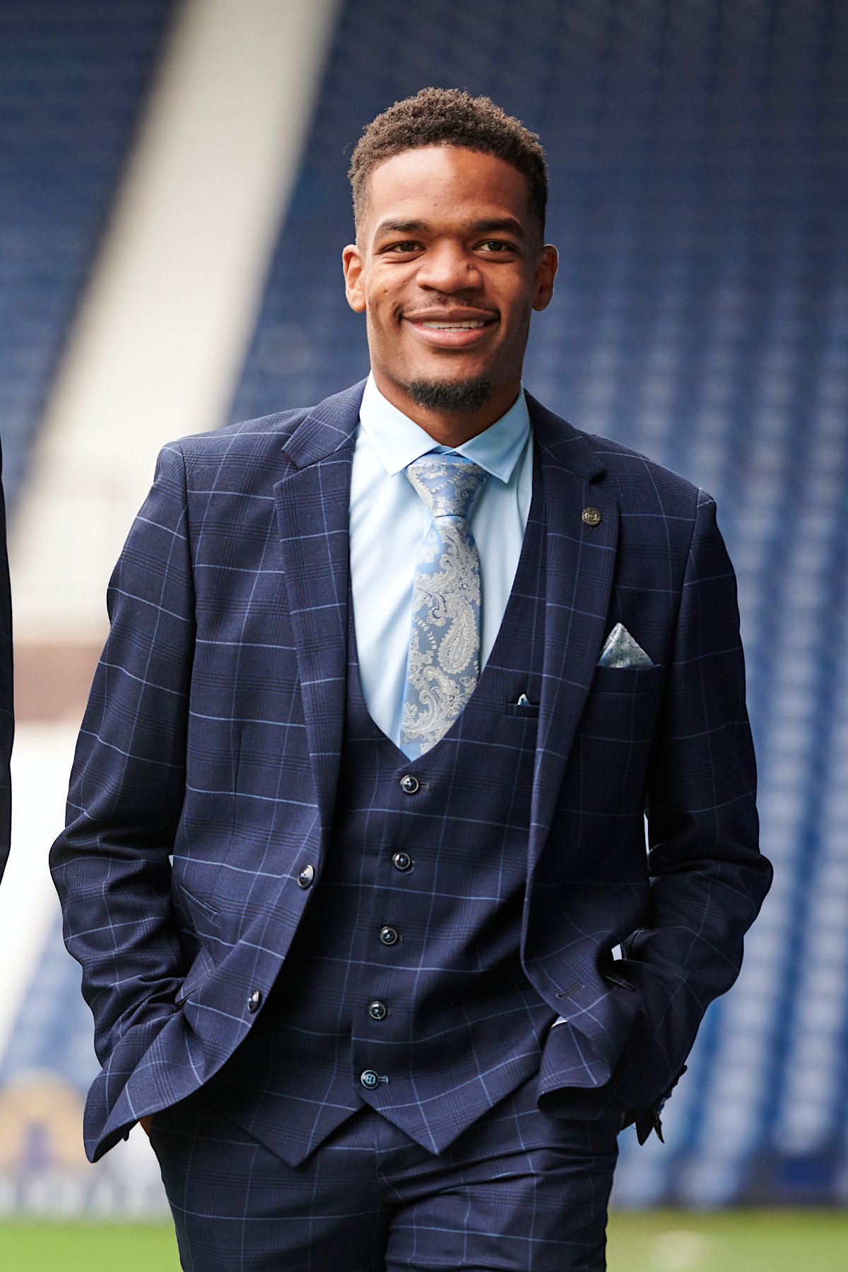 A man wearing the marcdarcy WBA Collection - Edinson Navy & Sky Check Suit, with a light blue shirt and paisley tie, smiles with hands in his pockets in front of blurred West Bromwich Albion F.C. stadium seats.
