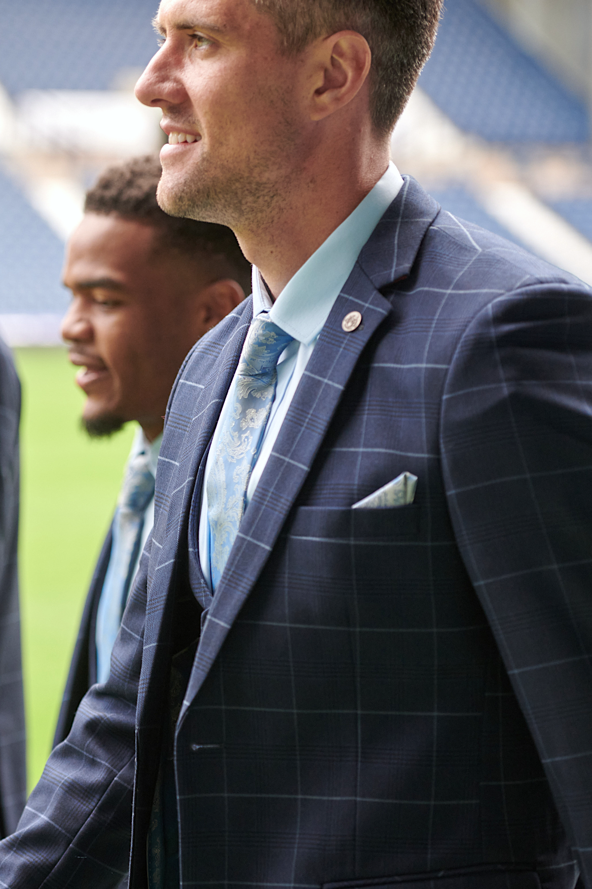 Two men in marcdarcys The WBA Collection - Edinson Navy & Sky Check Suits with light blue ties and pocket squares stand side by side, smiling, at a West Bromwich Albion F.C. stadium. The focus is on the man in the foreground.