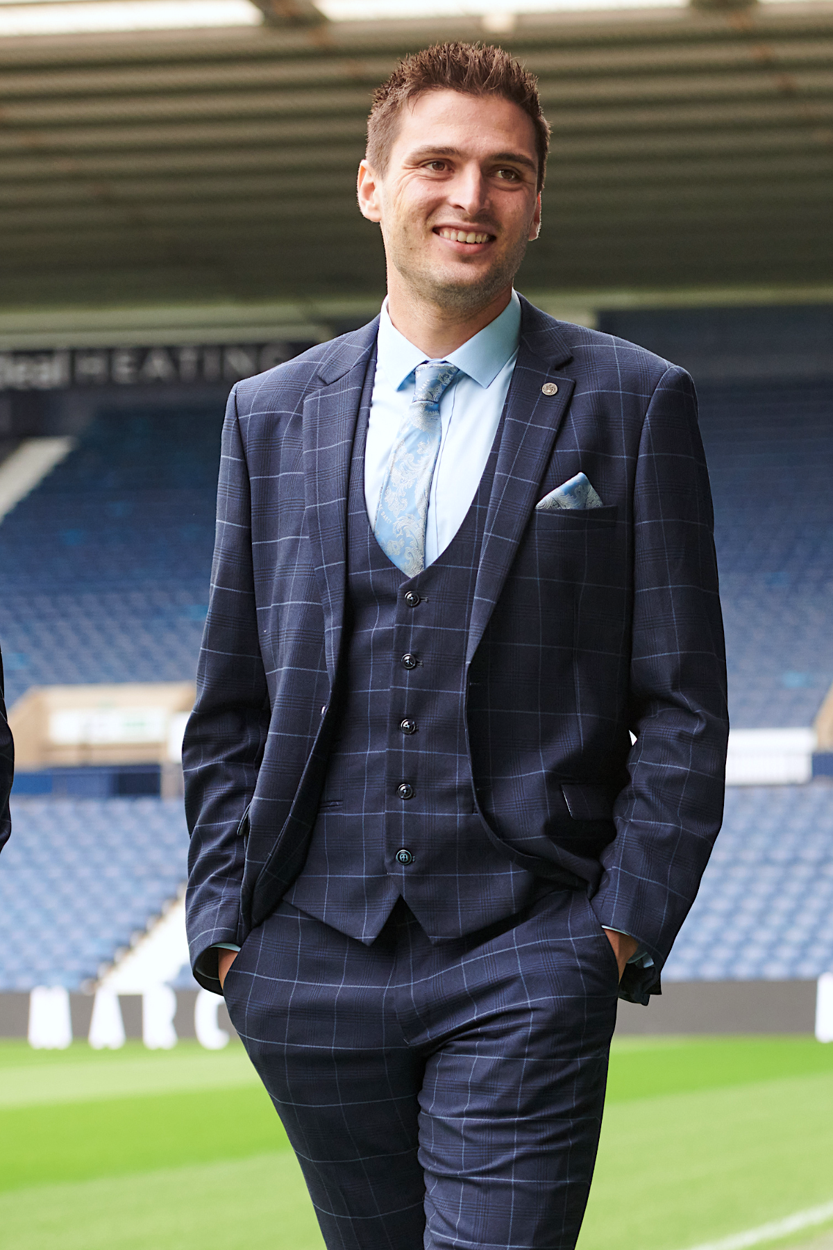 A man in the marcdarcy WBA Collection - Edinson Navy & Sky Check Suit stands on a football field, smiling with hands in pockets. Stadium seats and the roof at West Bromwich Albion F.C. are visible behind him.