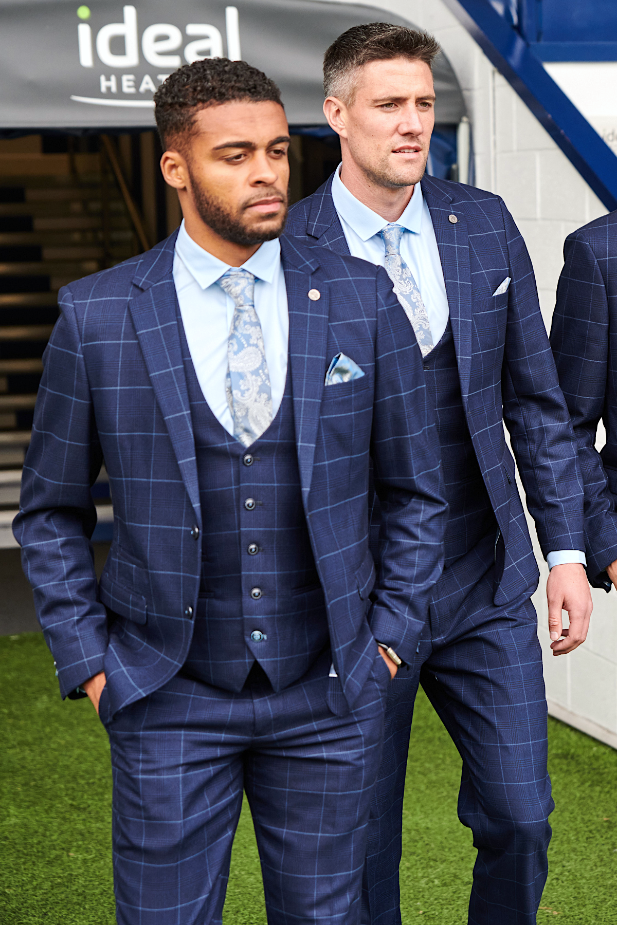 Two men wear marcdarcy's WBA Collection - Edinson Navy & Sky Check three-piece suits, paired with light blue shirts and patterned ties, as they walk forward on green turf.