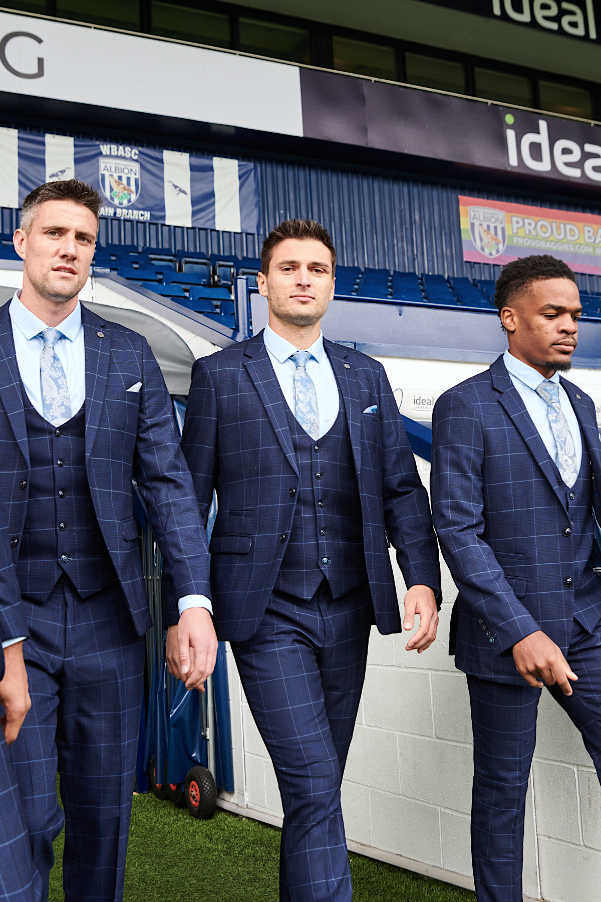 Three men in marcdarcy The WBA Collection - Edinson Navy & Sky Check Suits with light blue ties walk on a sports field, with West Bromwich Albion F.C. stadium seating and signage in the background.