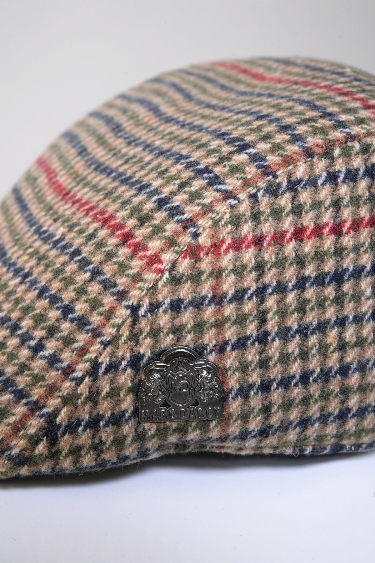 A close-up of the Marc Darcy EDWARD flat cap in olive green check tweed, inspired by the Peaky Blinders style, with beige, blue, red, and green stripes and a metallic Marc Darcy logo badge on the side.