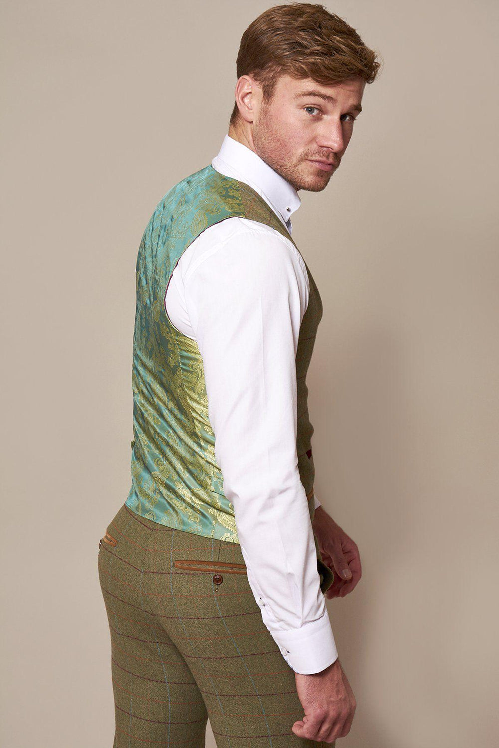 A man with short light brown hair wears a white shirt and brown plaid trousers, turning to reveal the marcdarcy ELLIS Moss Green Check Tweed Waistcoat, its slim fit evoking classic Peaky Blinders style as he looks over his shoulder at the camera.