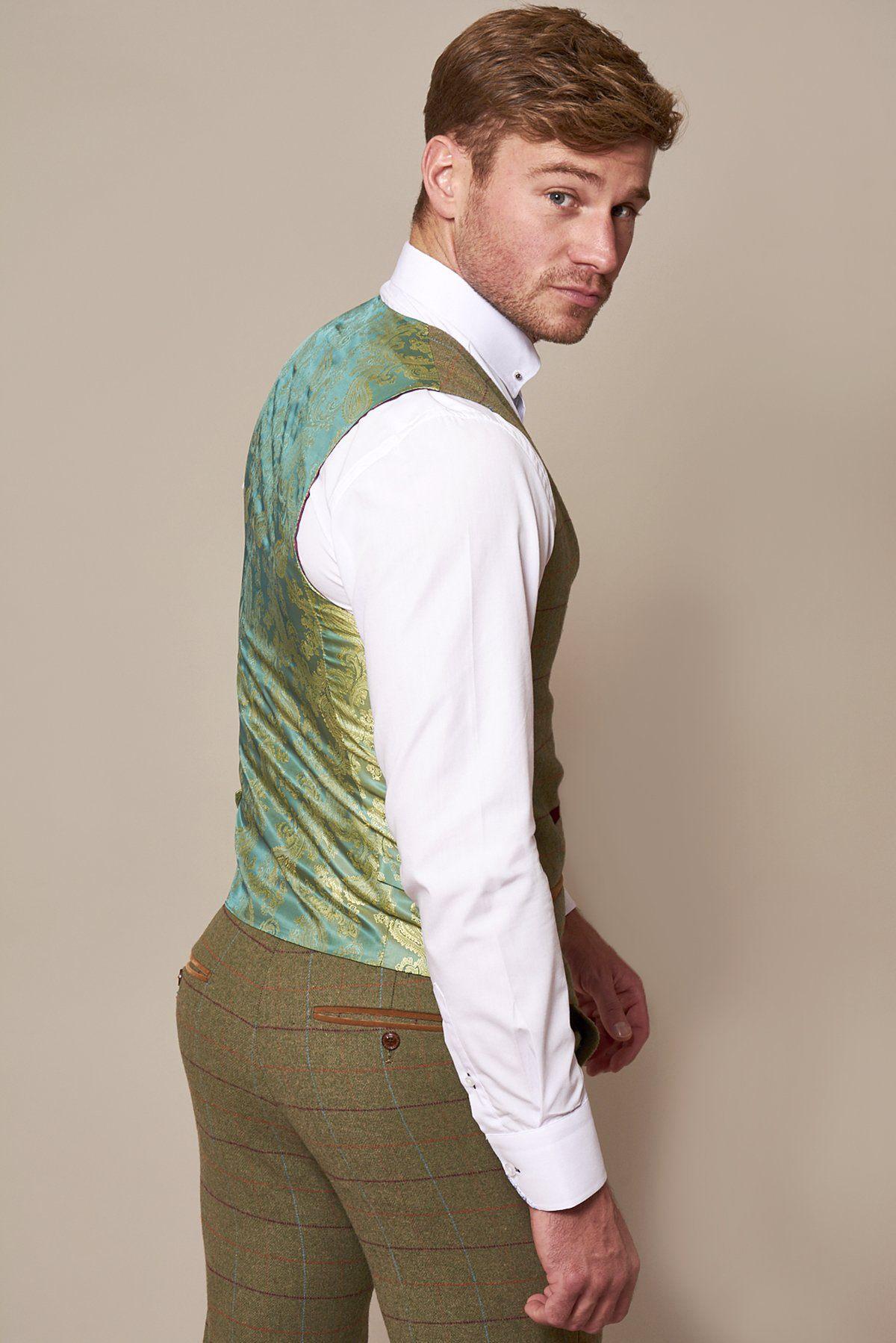 A man with short light brown hair wears a white shirt and brown plaid trousers, turning to reveal the marcdarcy ELLIS Moss Green Check Tweed Waistcoat, its slim fit evoking classic Peaky Blinders style as he looks over his shoulder at the camera.