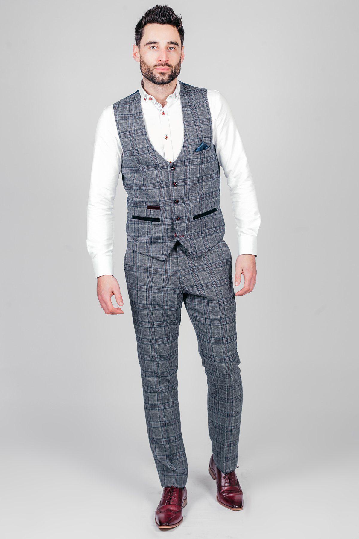 A man with short dark hair and a beard stands against a plain background wearing a marcdarcy ENZO Blue Grey Check Tweed Waistcoat, matching check trousers, a white shirt, and burgundy dress shoes for a Peaky Blinders-inspired look.