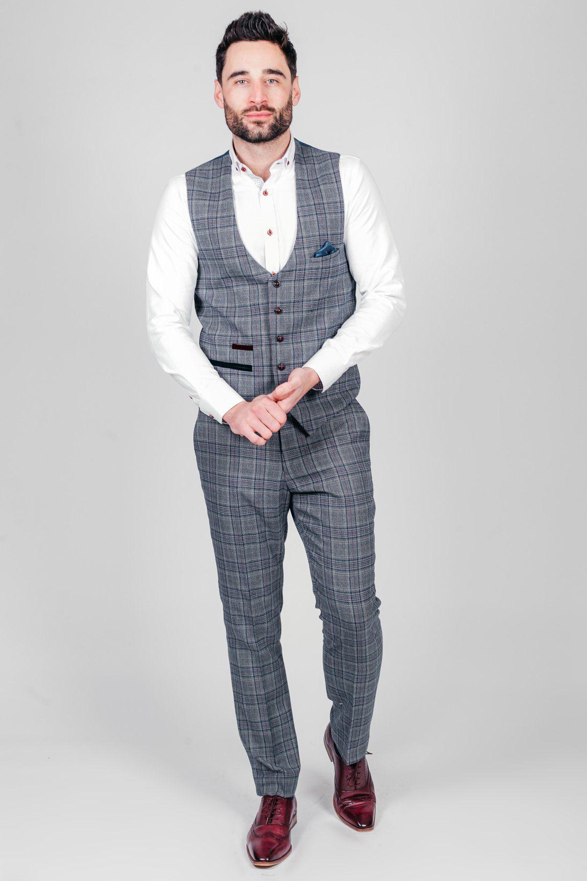 A man with short dark hair and a beard stands against a light gray background in a marcdarcy ENZO Blue Grey Check Tweed Single Breasted Waistcoat, white shirt, and burgundy dress shoes, hands clasped in front.