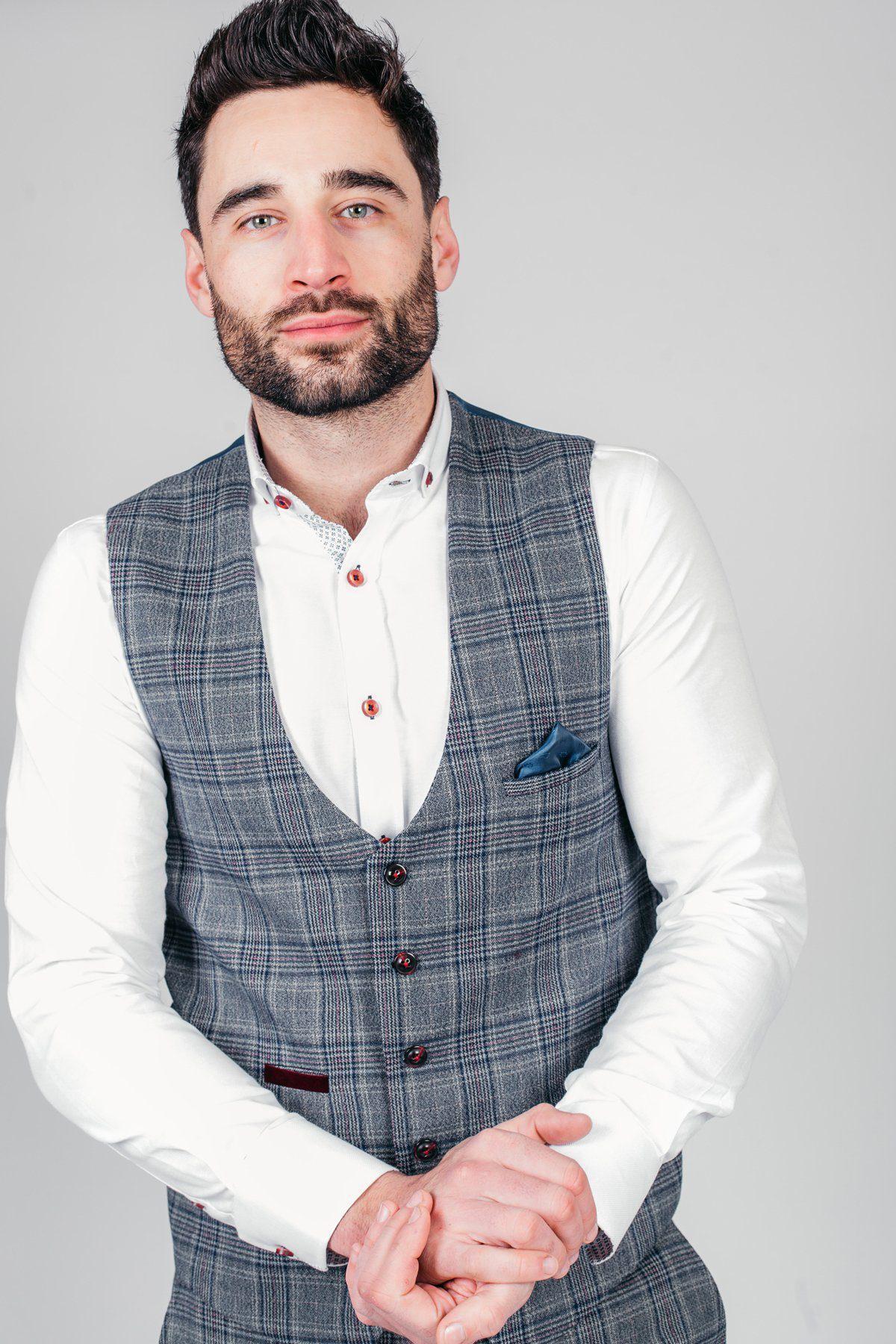 Man with dark hair and beard wears a white shirt and the marcdarcy ENZO Blue Grey Check Tweed Single Breasted Waistcoat with a blue pocket square, standing against a plain light gray background, looking at the camera neutrally.