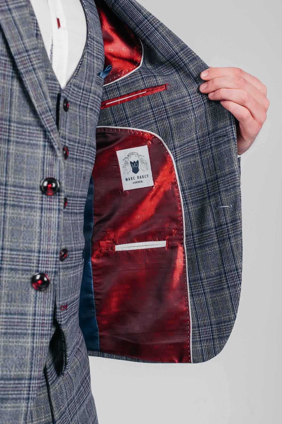 A person opens an ENZO - Blue Grey Tweed Check Blazer by marcdarcy, revealing its shiny red patterned lining and white marcdarcy label. The Peaky Blinders-inspired blazer features dark buttons and is styled over a white shirt.