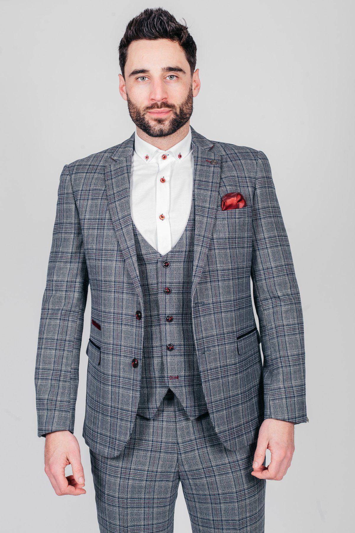 A man with dark hair and a beard wears marcdarcy’s ENZO - Blue Grey Tweed Check Blazer, paired with a white dress shirt with red buttons and a red pocket square, standing against a plain light background.
