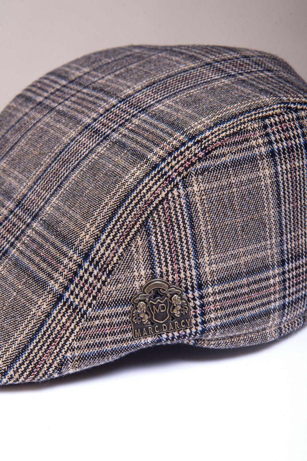 A close-up of the ENZO - Tan Tweed Flat Cap by Accessory, showcasing its Prince of Wales check pattern in brown and grey plaid with a metallic logo pin on the side.