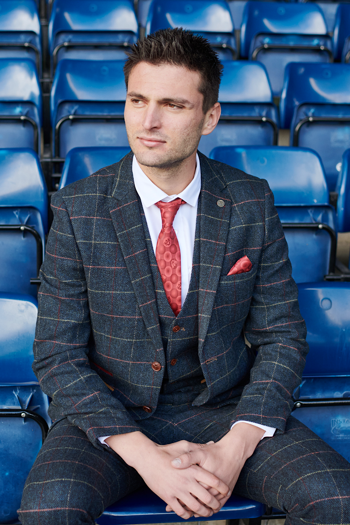 Wearing the marcdarcy WBA Collection - Eton Suit as worn by Okay Yokuşlu, a man in a slim fit blazer with a red tie and pocket square sits on blue stadium seats, looking left with hands on his knees.