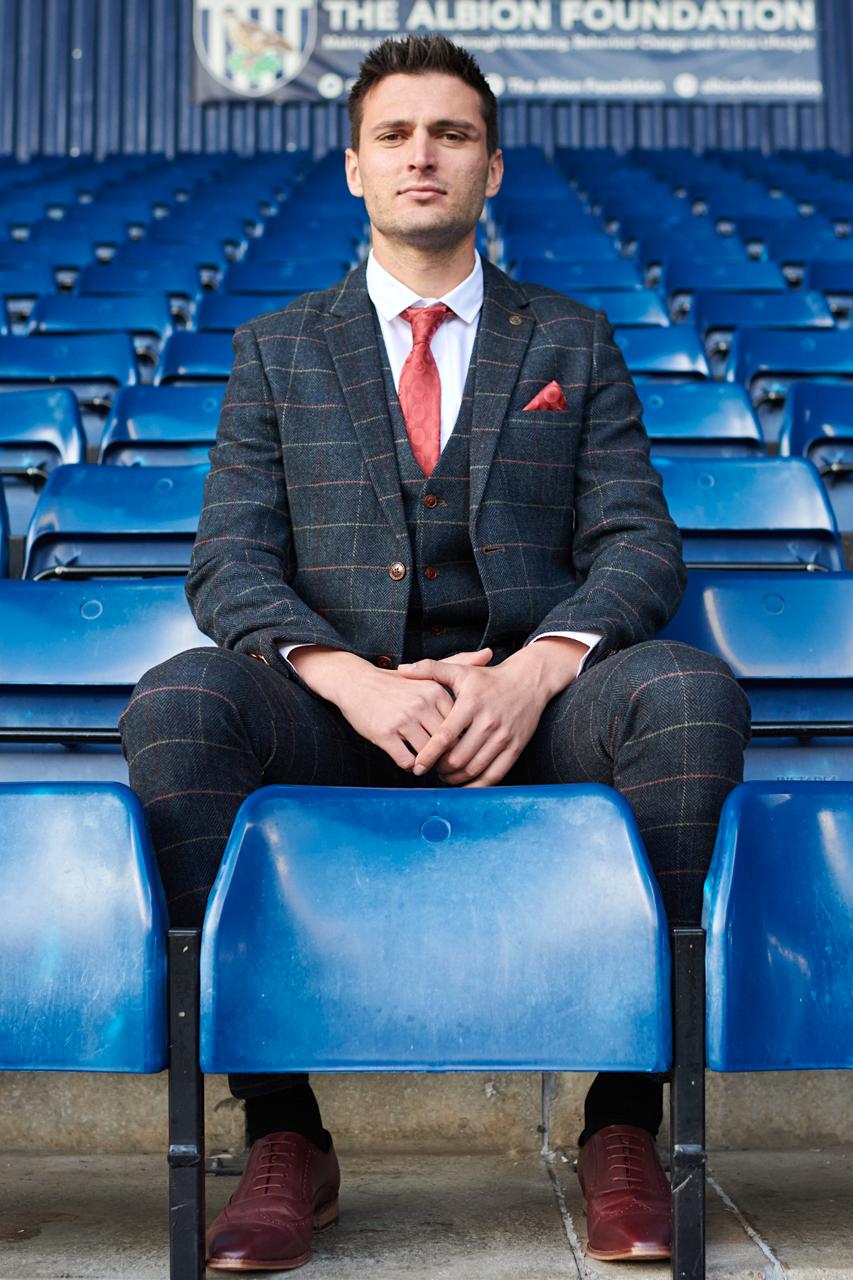 A man in The WBA Collection - Eton Suit by marcdarcy, as worn by Okay Yokuşlu, sits with hands on his knees in blue stadium seats, a sign for The Albion Foundation visible behind him.