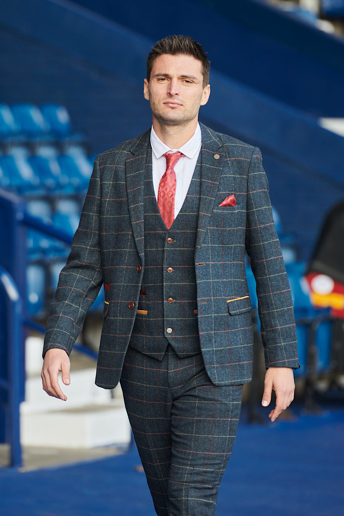 A man wears the marcdarcy WBA Collection - Eton Suit as worn by Okay Yokuşlu, with a red tie and pocket square, walking toward the camera in front of blue stadium seats.