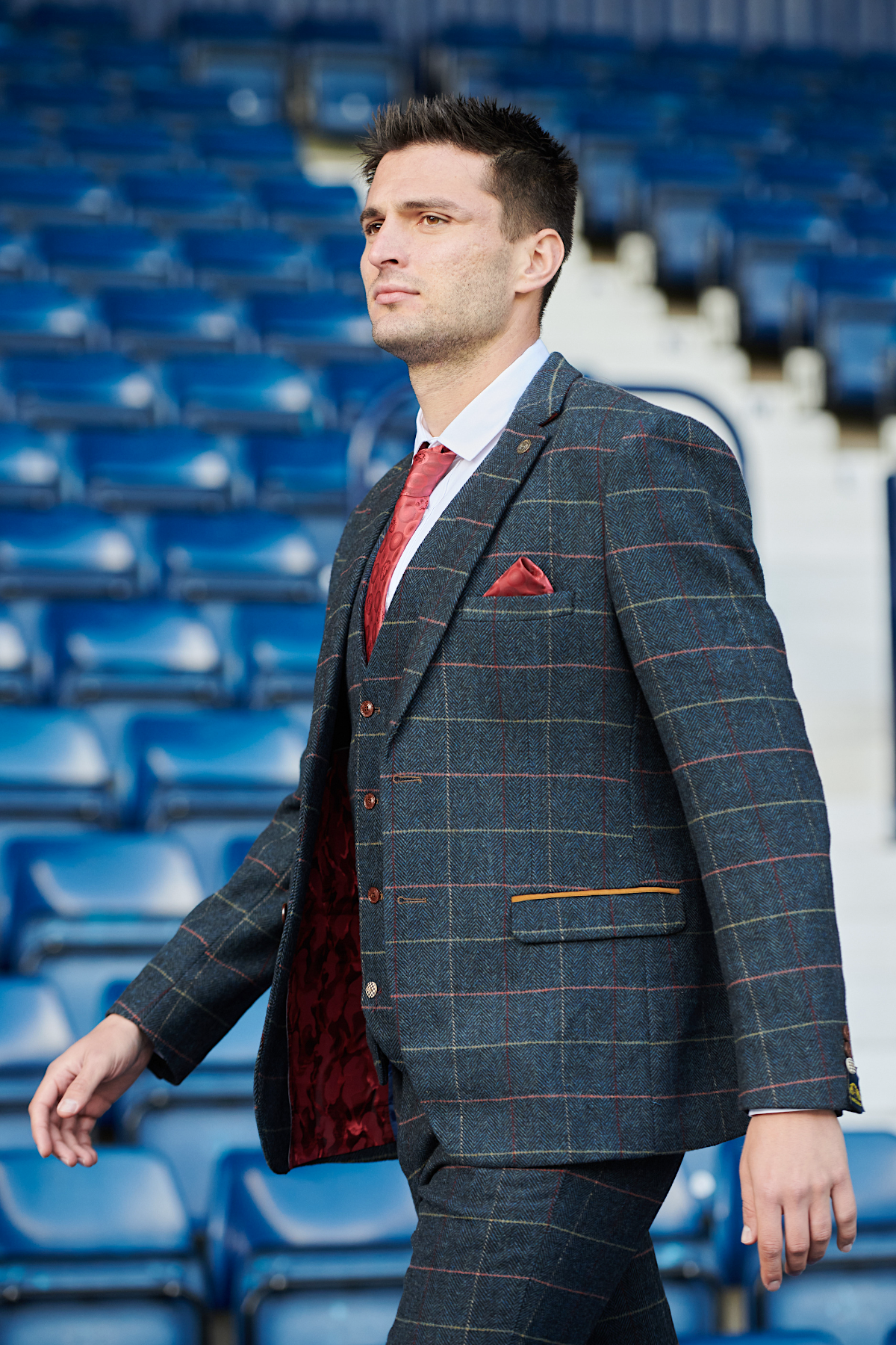 A man wearing The WBA Collection - Eton Suit by marcdarcy, as worn by Okay Yokuşlu, paired with a red tie and pocket square, walks confidently through a stadium lined with empty blue seats.