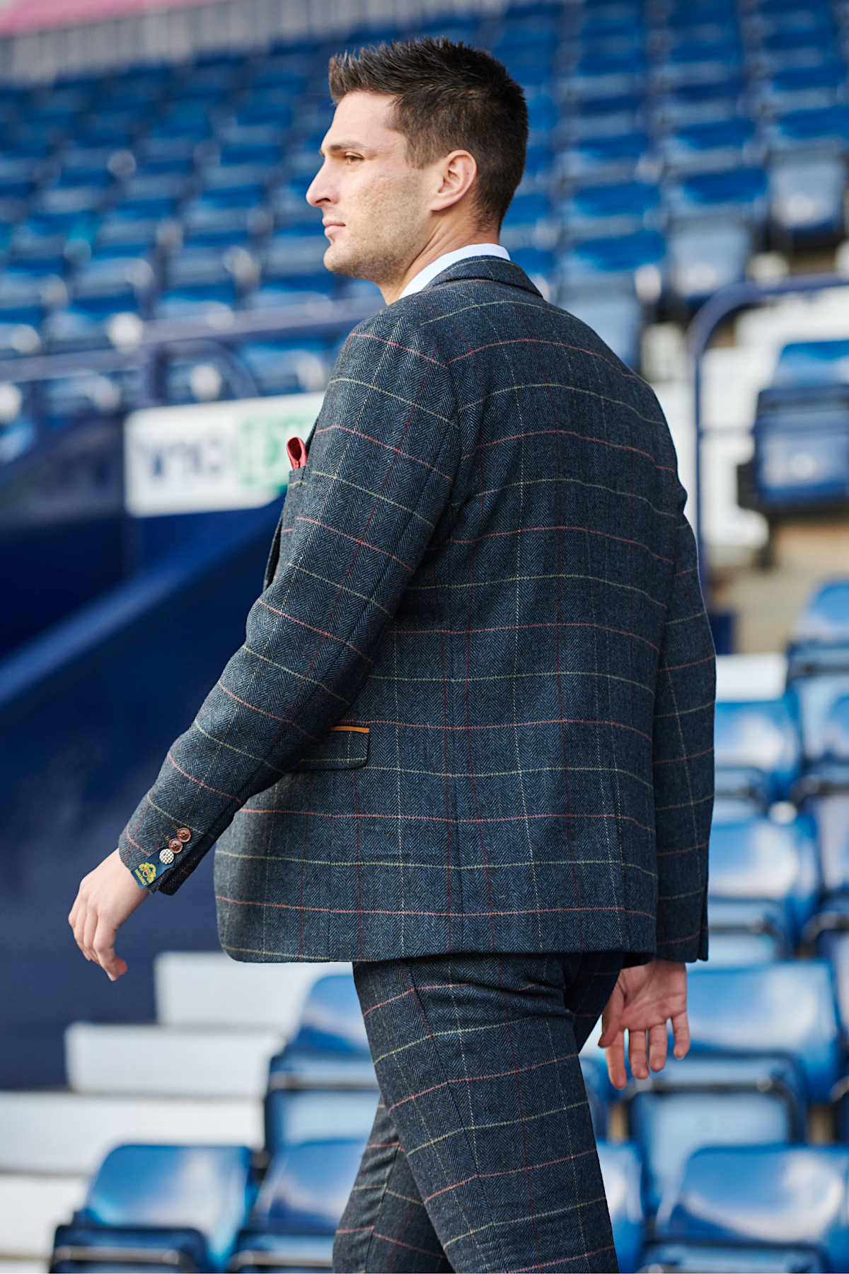 A man in The WBA Collection – Eton Suit As Worn By Okay Yokuşlu from marcdarcy walks confidently past empty blue stadium seats, facing left.