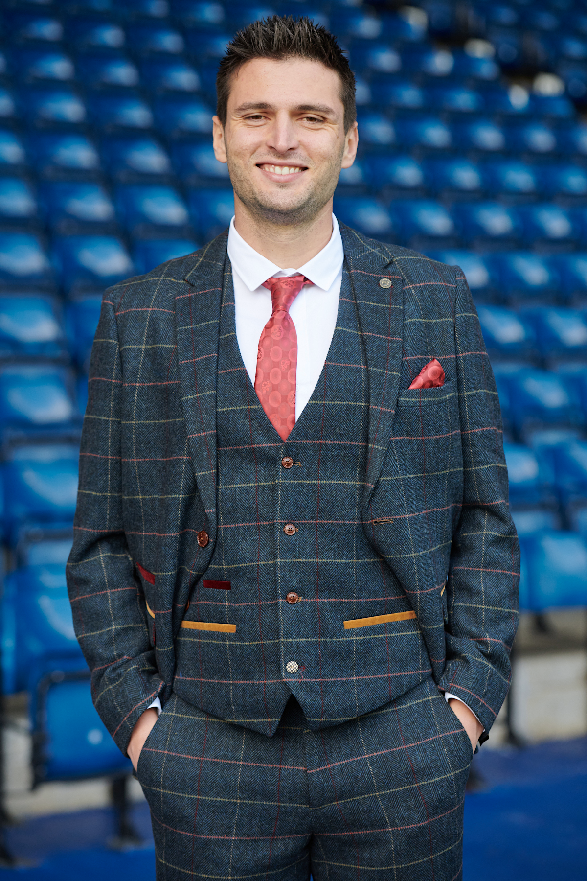 A man in The WBA Collection - Eton Suit by marcdarcy, as worn by Okay Yokuşlu, stands smiling with hands in his pockets in front of empty blue stadium seats, wearing a red tie and pocket square.