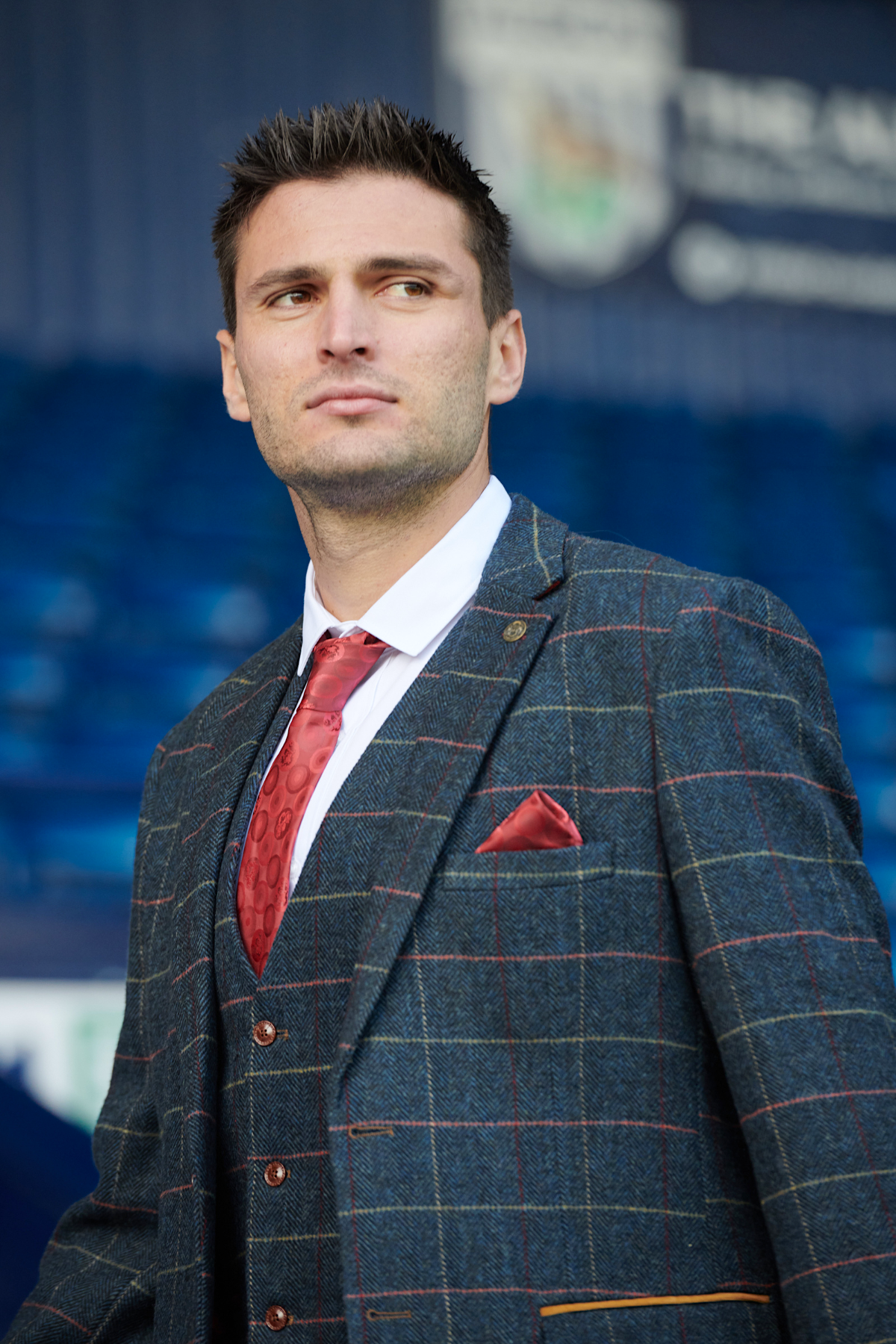 A man wearing the marcdarcy WBA Collection - Eton Suit as worn by Okay Yokuşlu, with a red tie and pocket square, stands thoughtfully in a stadium with blue seats and a blurred sports emblem in the background.